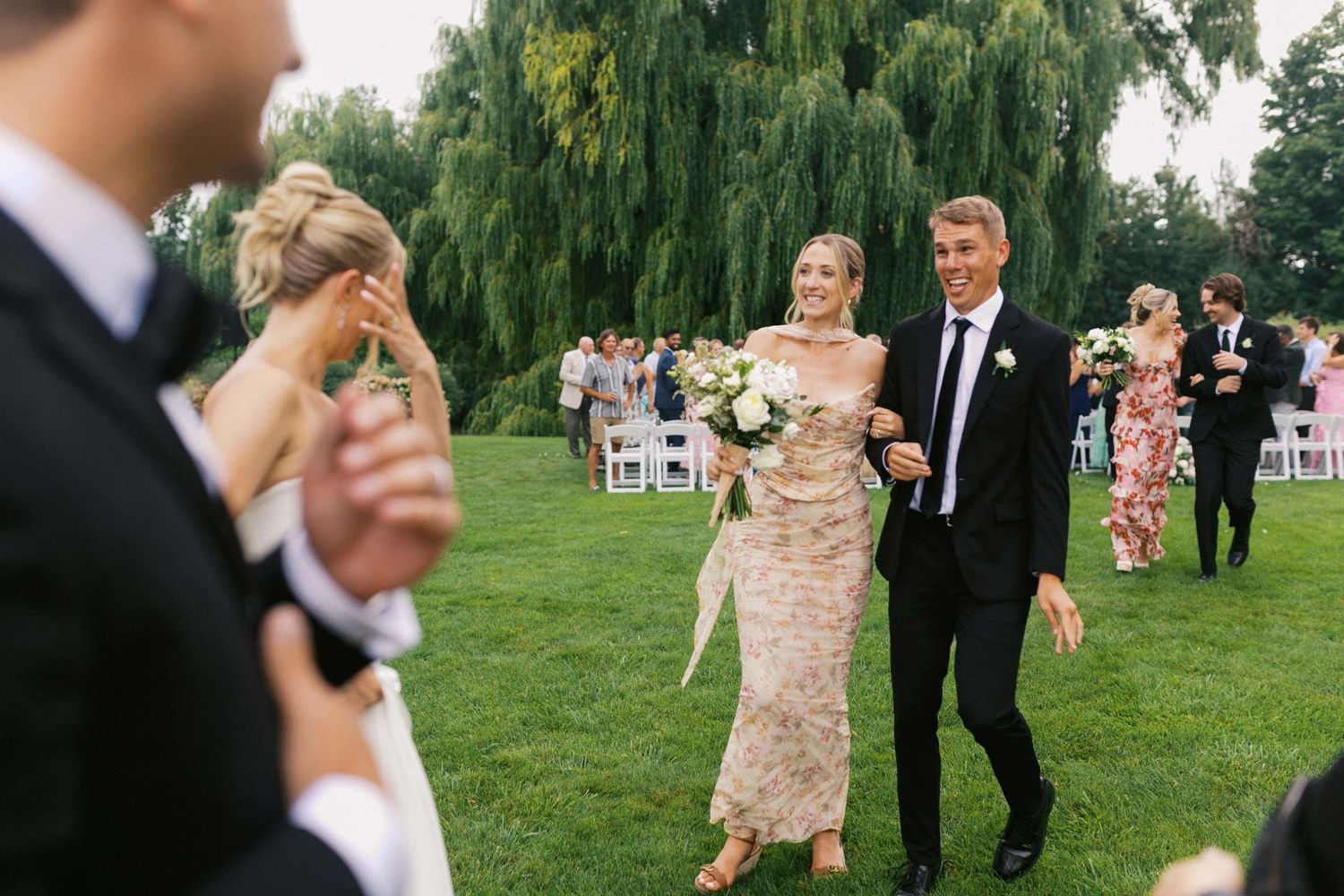 Wedding friends show emotion on a lush green lawn beneath weeping willow trees after an elegant outdoor ceremony.