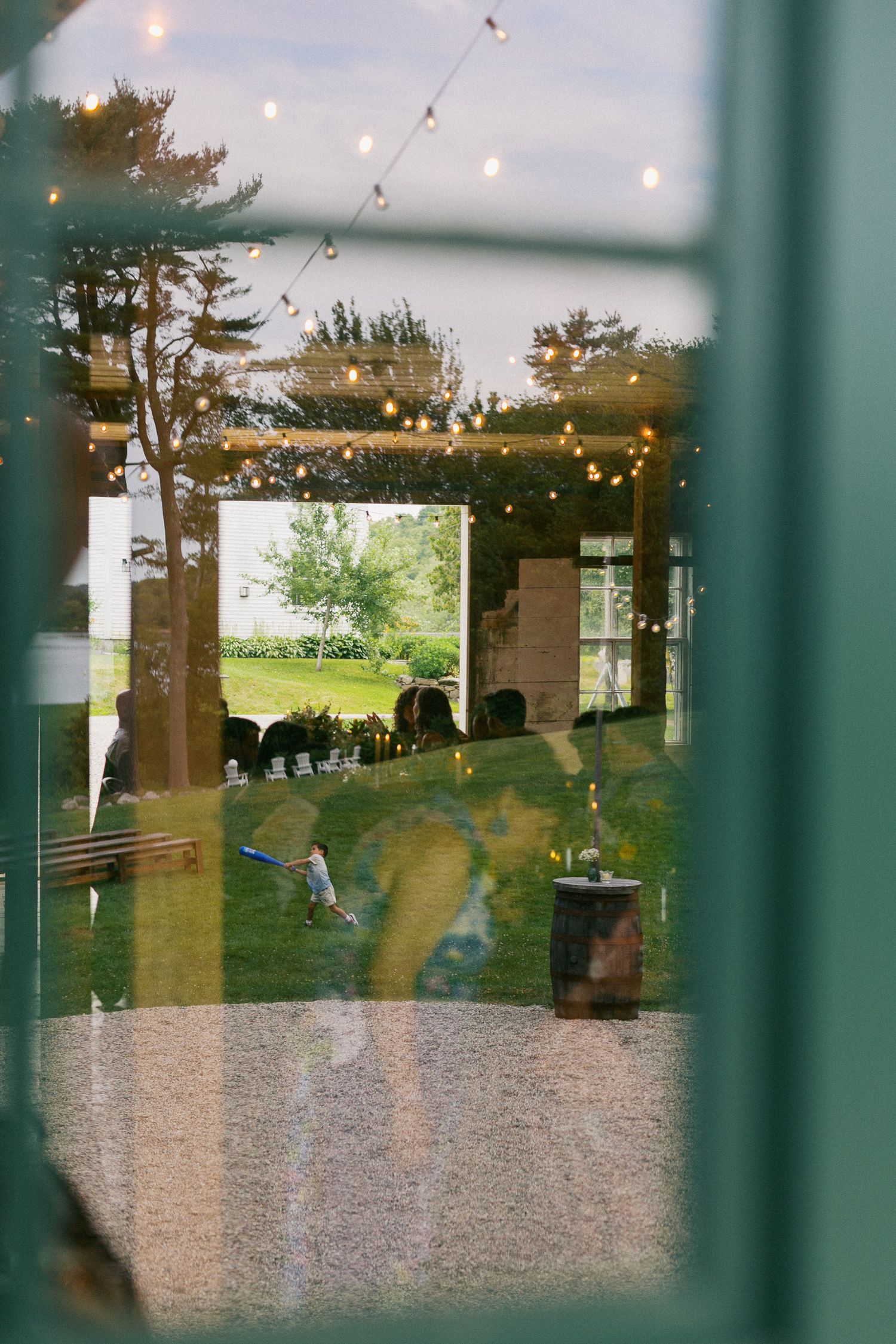 Outdoor garden venue with string lights, pergola, and lush greenery viewed through a window reflection.