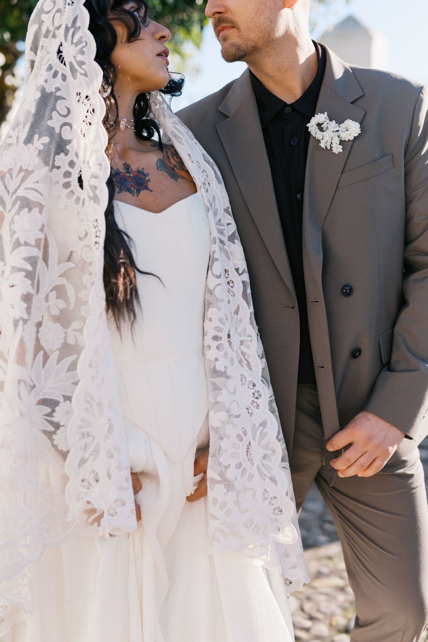 Interracial couple in wedding attire, bride in lace veil and white gown with tattoos, groom in gray suit with boutonniere.