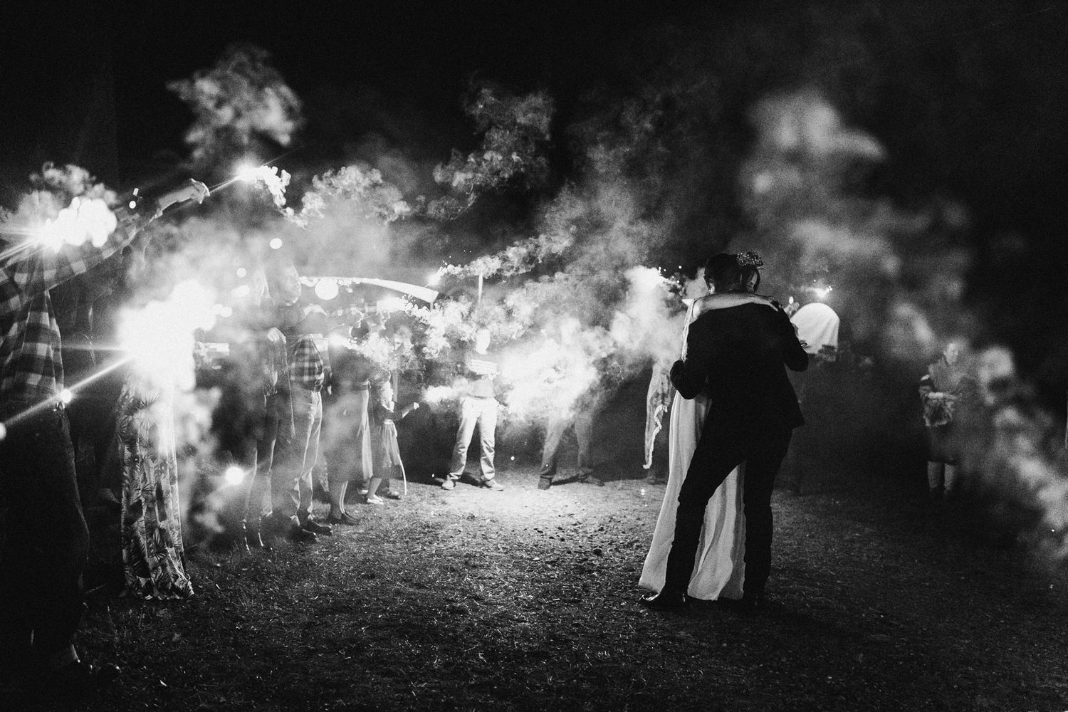 Black and white wedding couple kissing under sparklers held by guests at night.