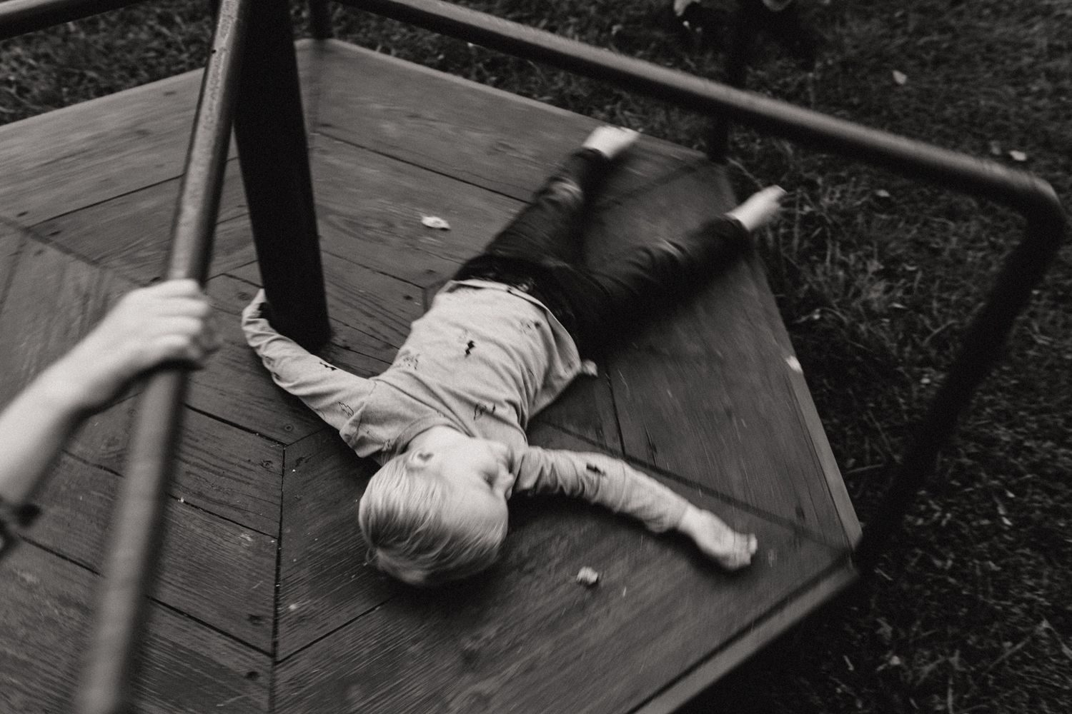 Black and white photo of a child lying on a wooden merry-go-round at a playground.