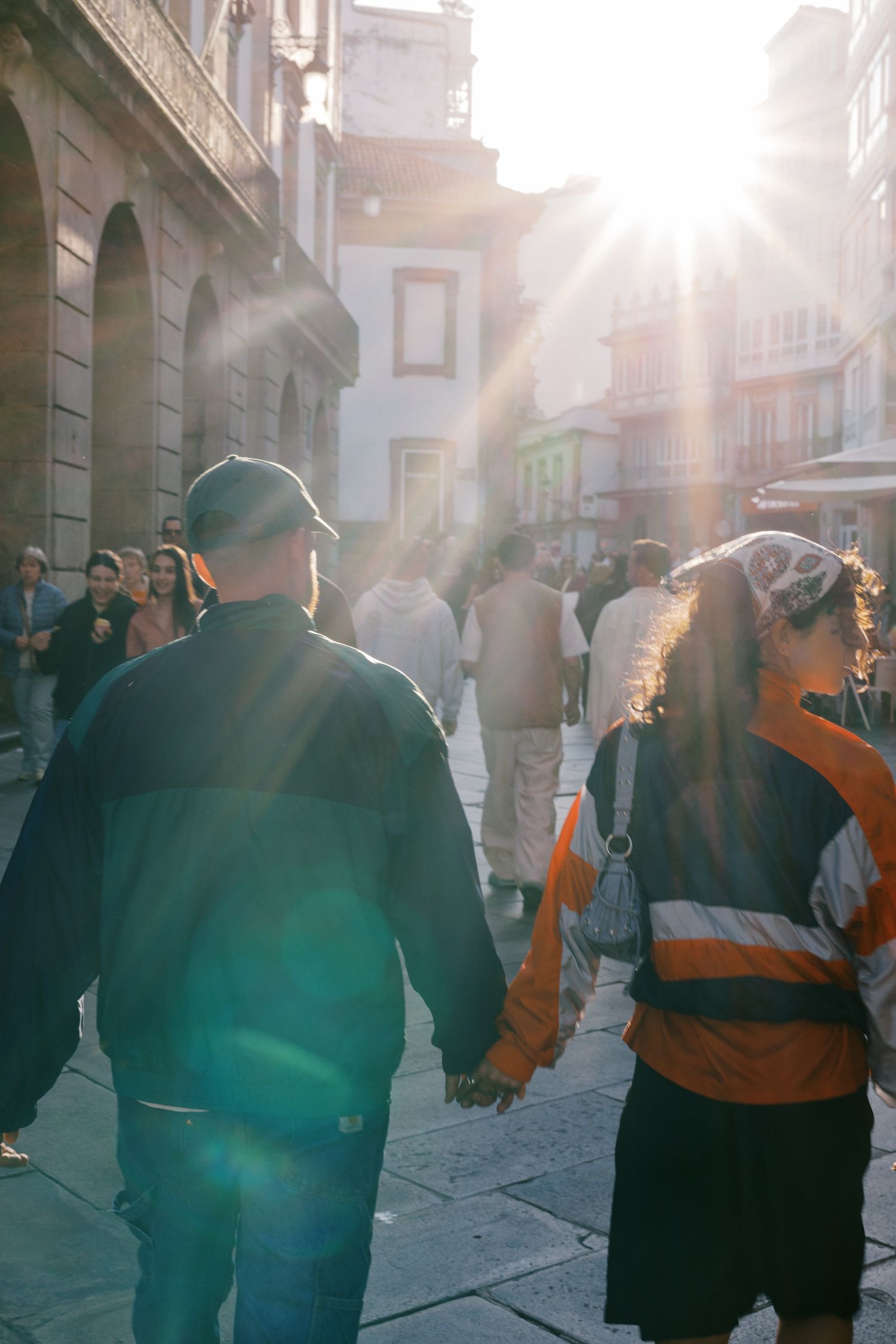 Couple holding hands walking through a sunlit cobblestone street in a European city.