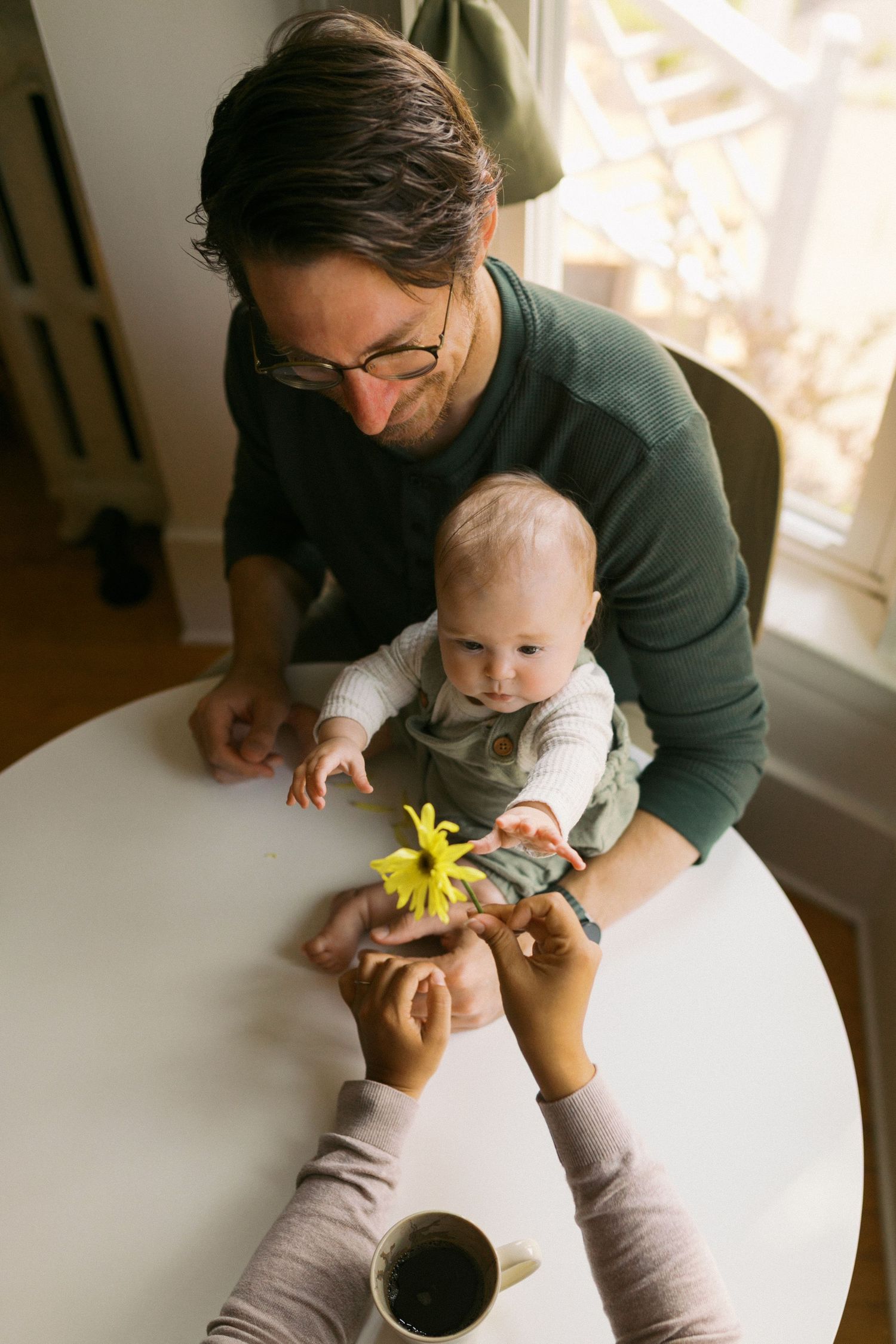 Father holds baby at white table while mother offers a bright yellow flower in warm home setting.