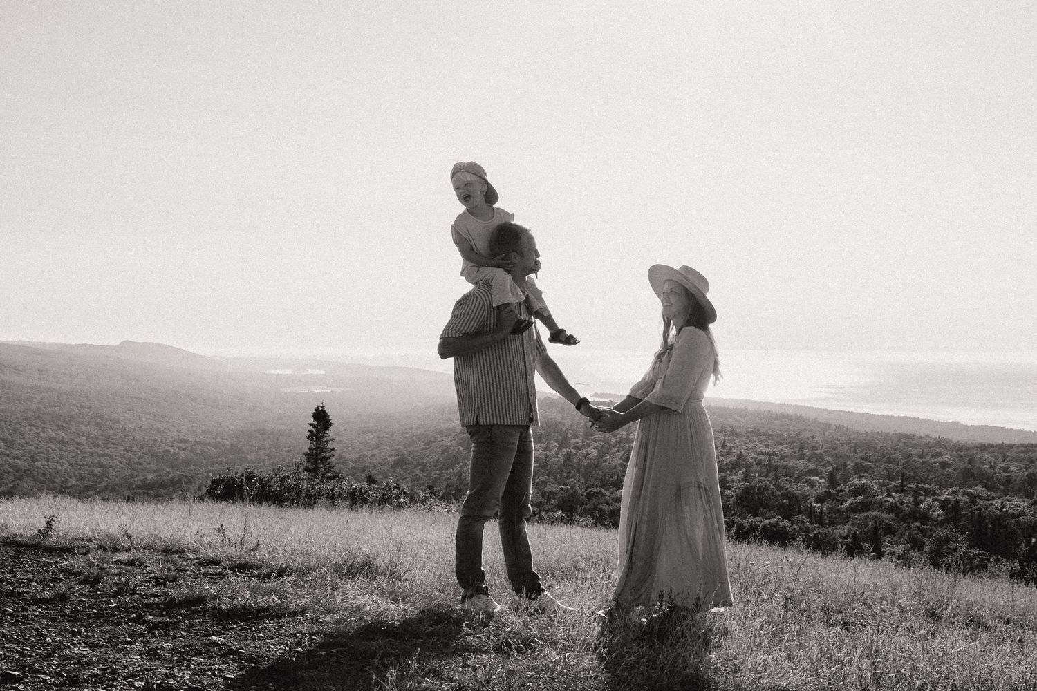 Black and white family portrait with child on fathers shoulders holding mothers hand on scenic hilltop.
