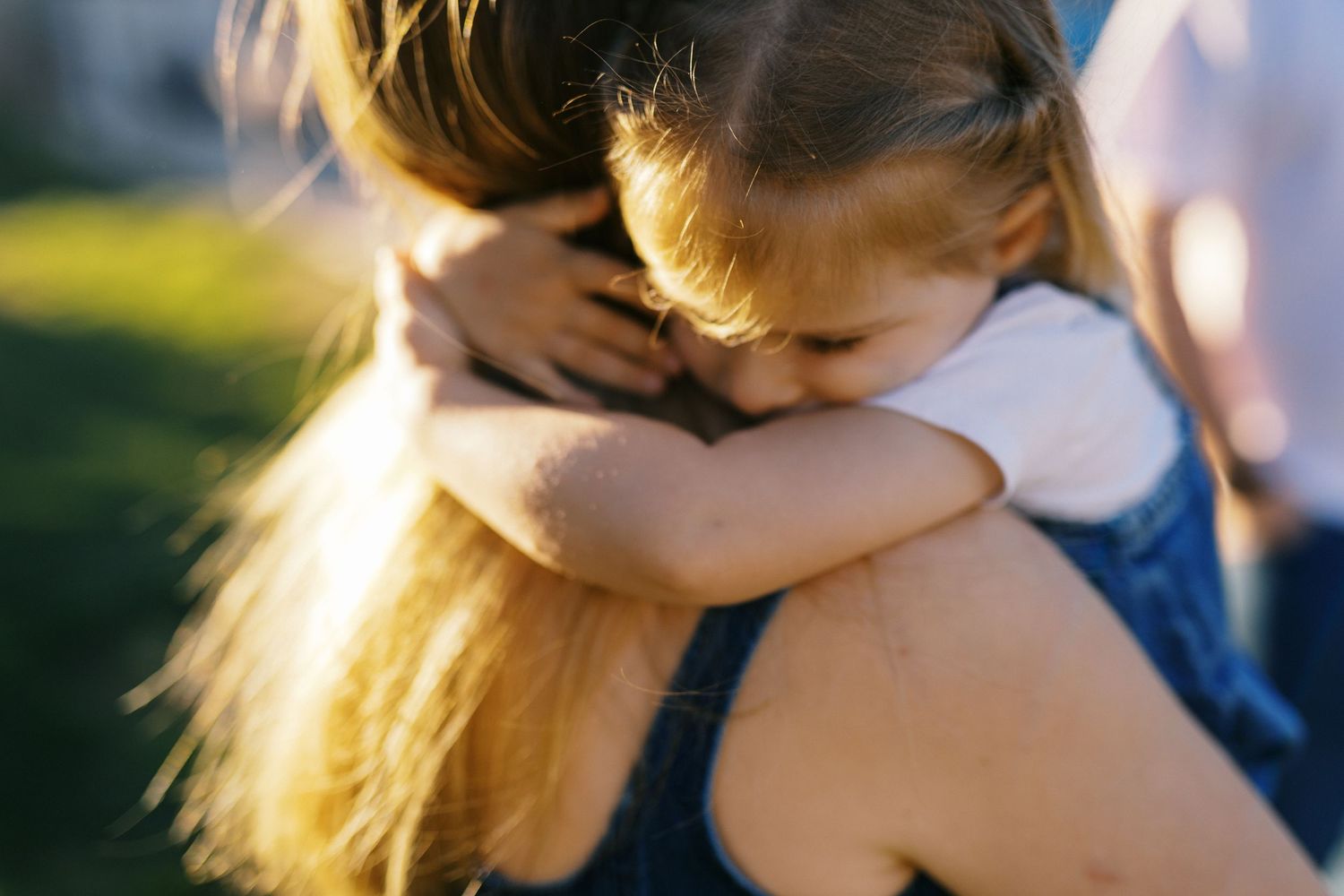 A young girl hugging her mother tightly outdoors in warm golden sunlight, showing a tender parent-child bond.