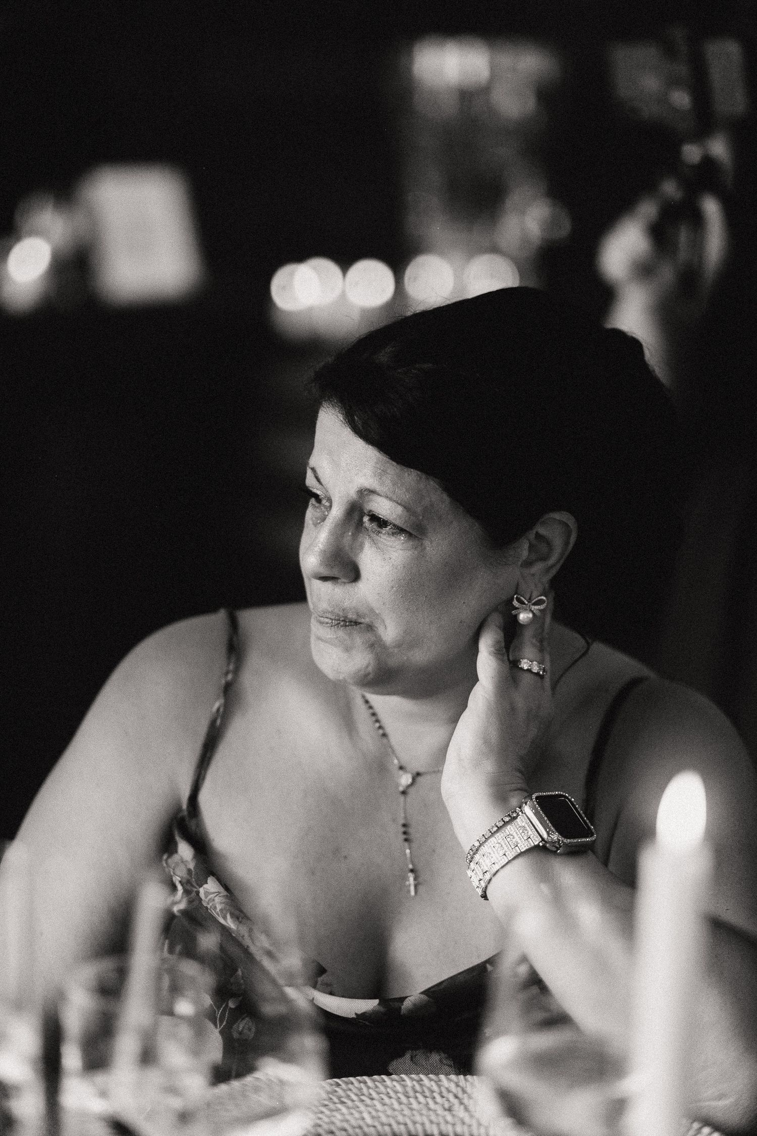 Black and white photo of an emotional mother of the bride at a candlelit reception, wearing jewelry and a smartwatch.