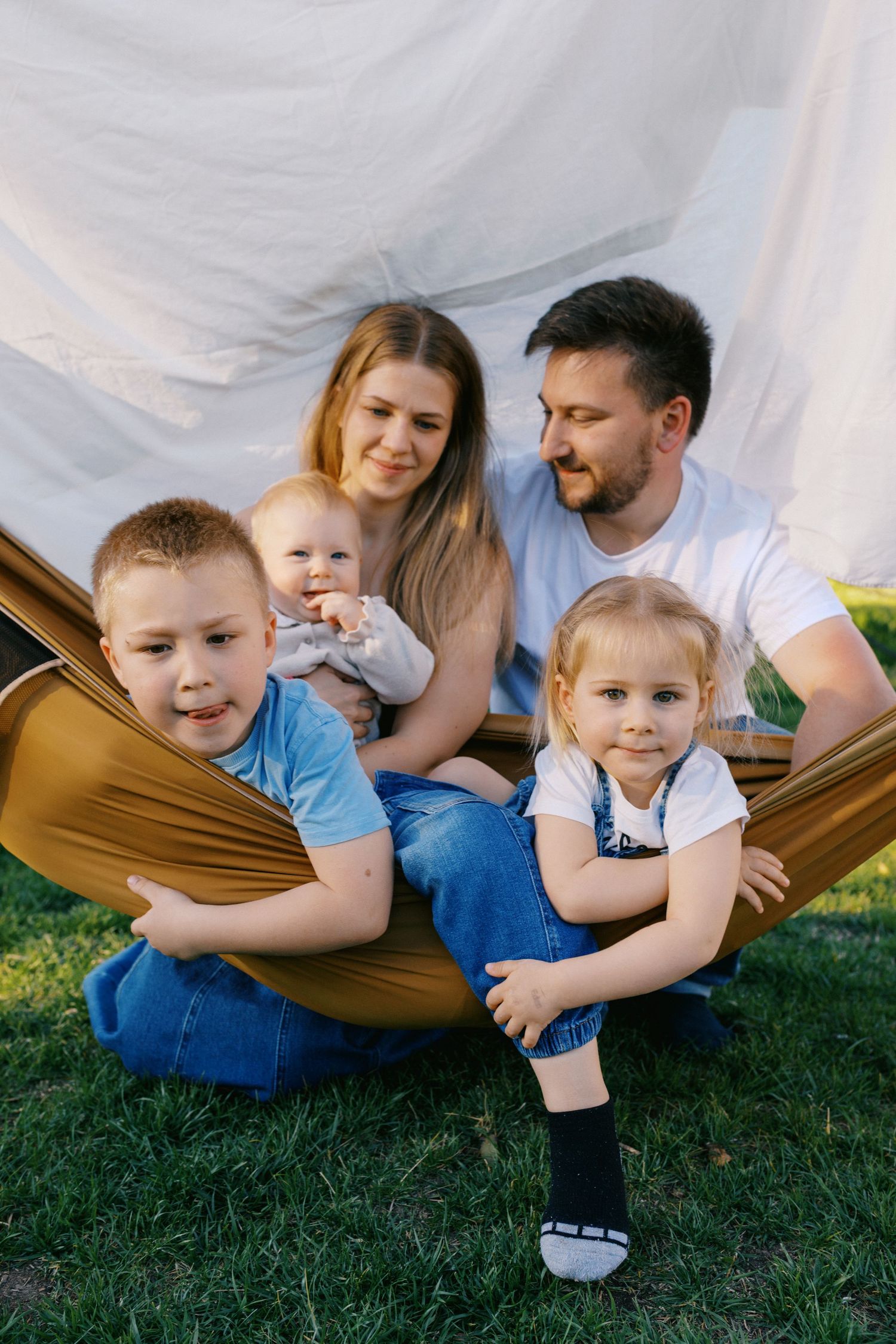 Happy family of five sitting together in a hammock outdoors on green grass with a white backdrop.
