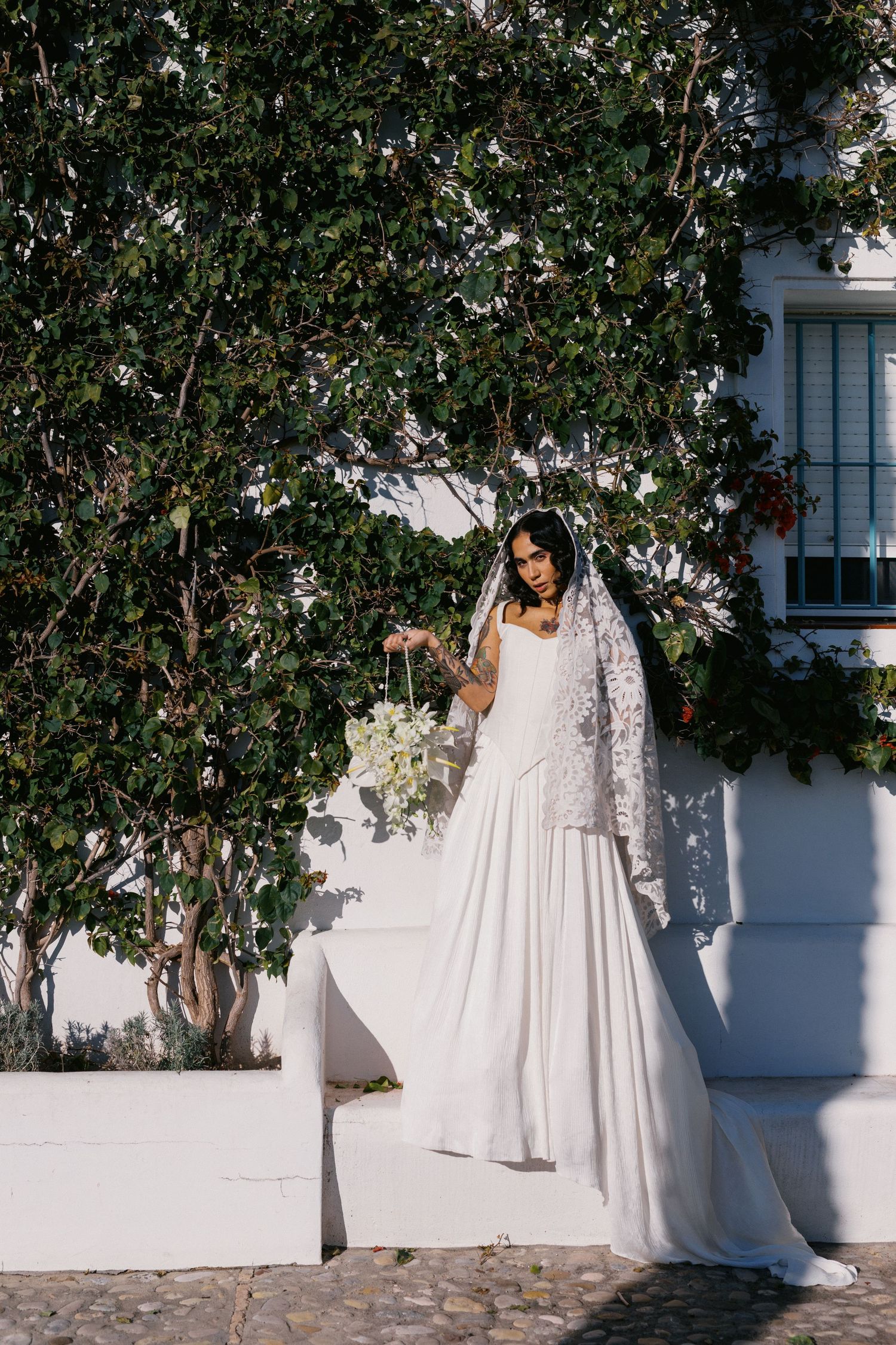 Bride in white lace veil and gown holds bouquet beside a white wall with lush green trees.
