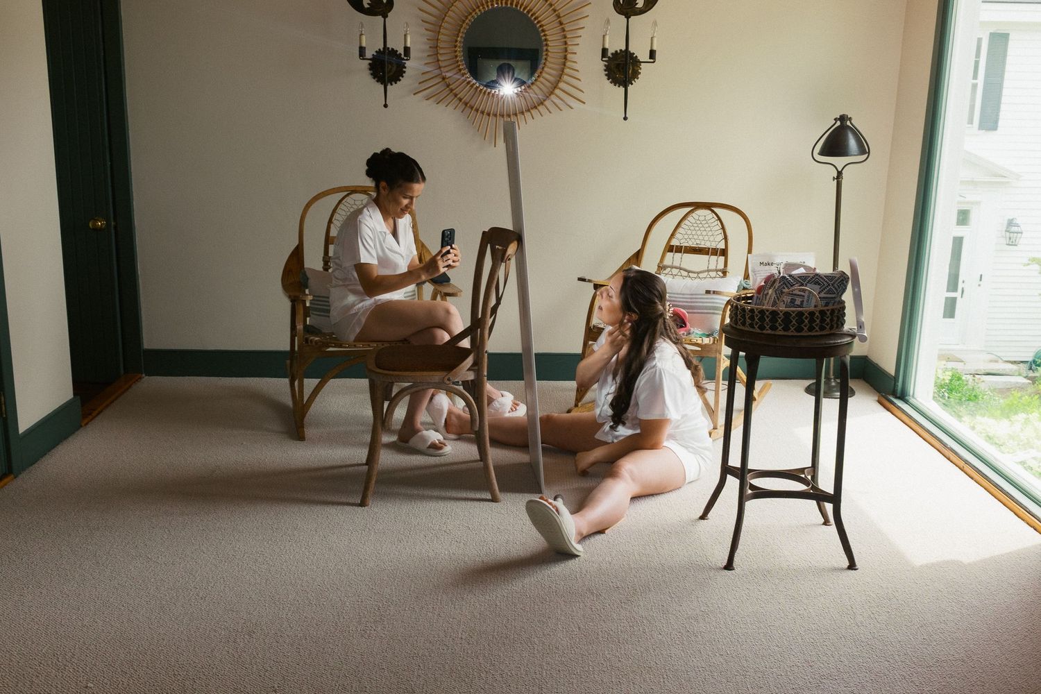 Two women relaxing in a sunlit room with rattan chairs, one sitting on the floor doing her makeup.