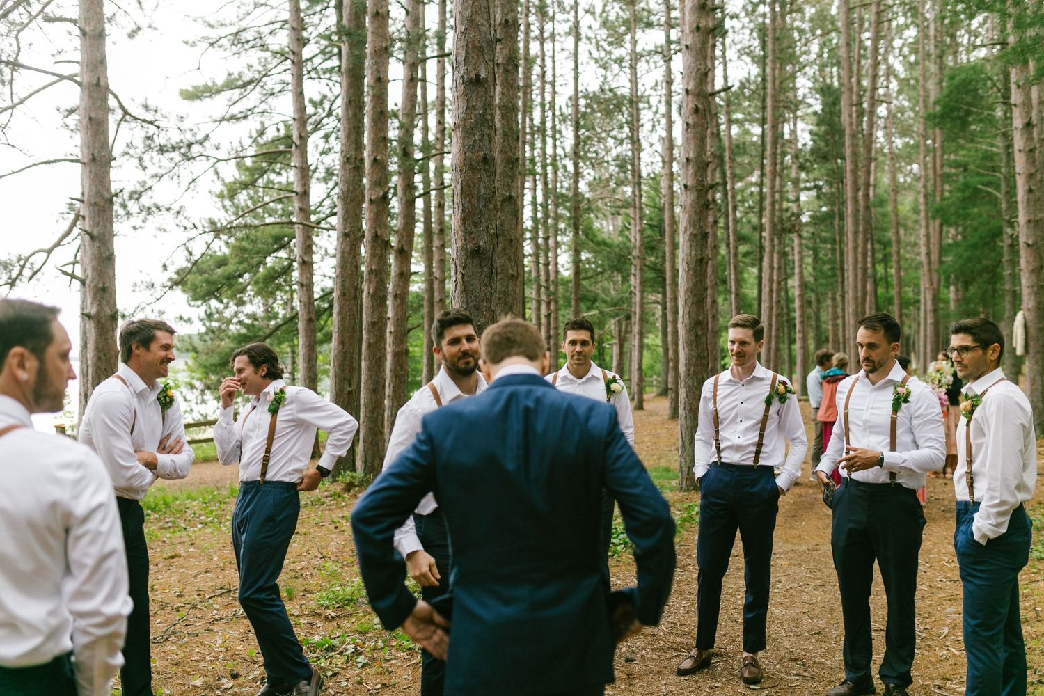 A group in white shirts and suspenders gather in a circle among tall pine trees during an outdoor event.