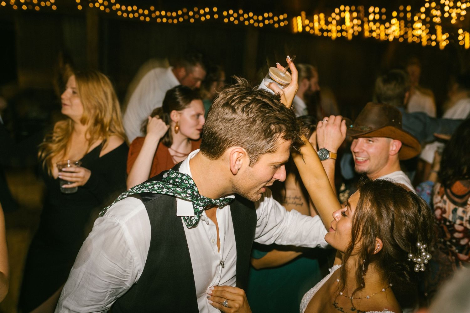 Wedding guests dancing and celebrating on a dimly lit dance floor decorated with string lights above.