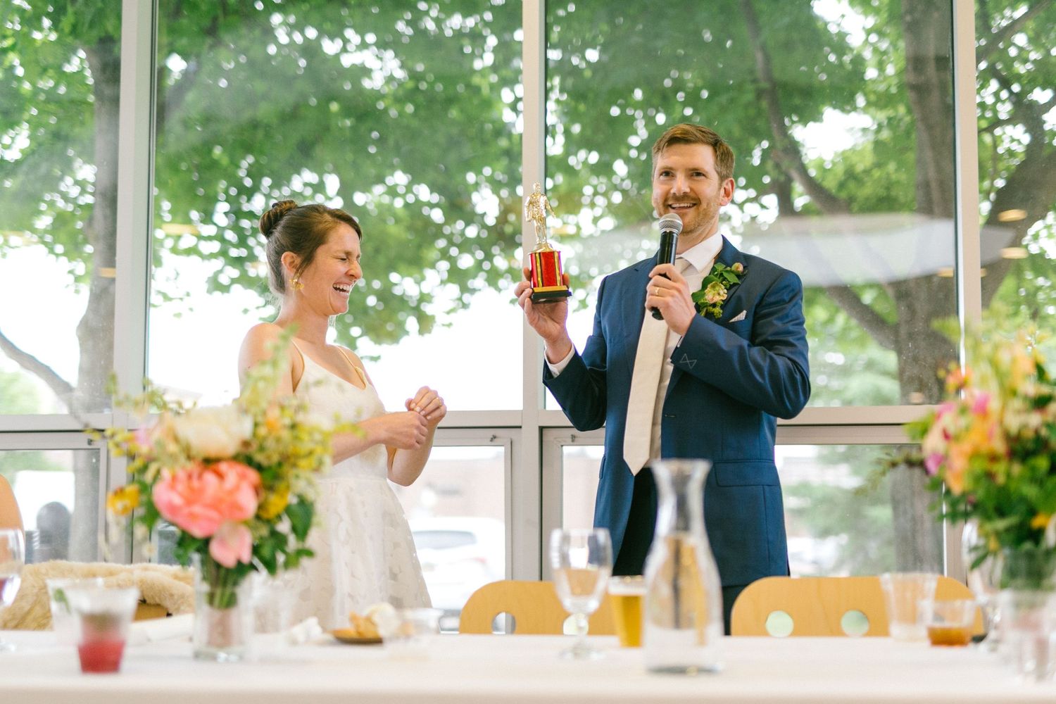 A wedding celebration scene with toasting in a sunlit glass-walled venue decorated with flowers.