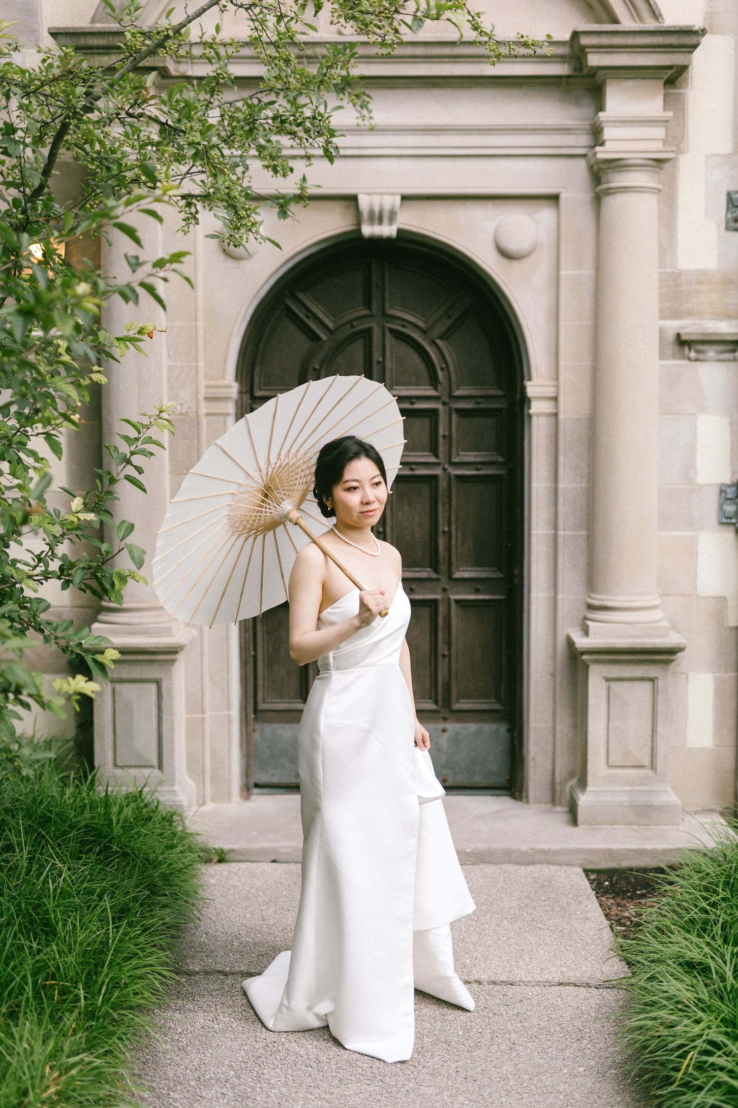 Bride in white gown holds parasol umbrella outside stone church entrance.