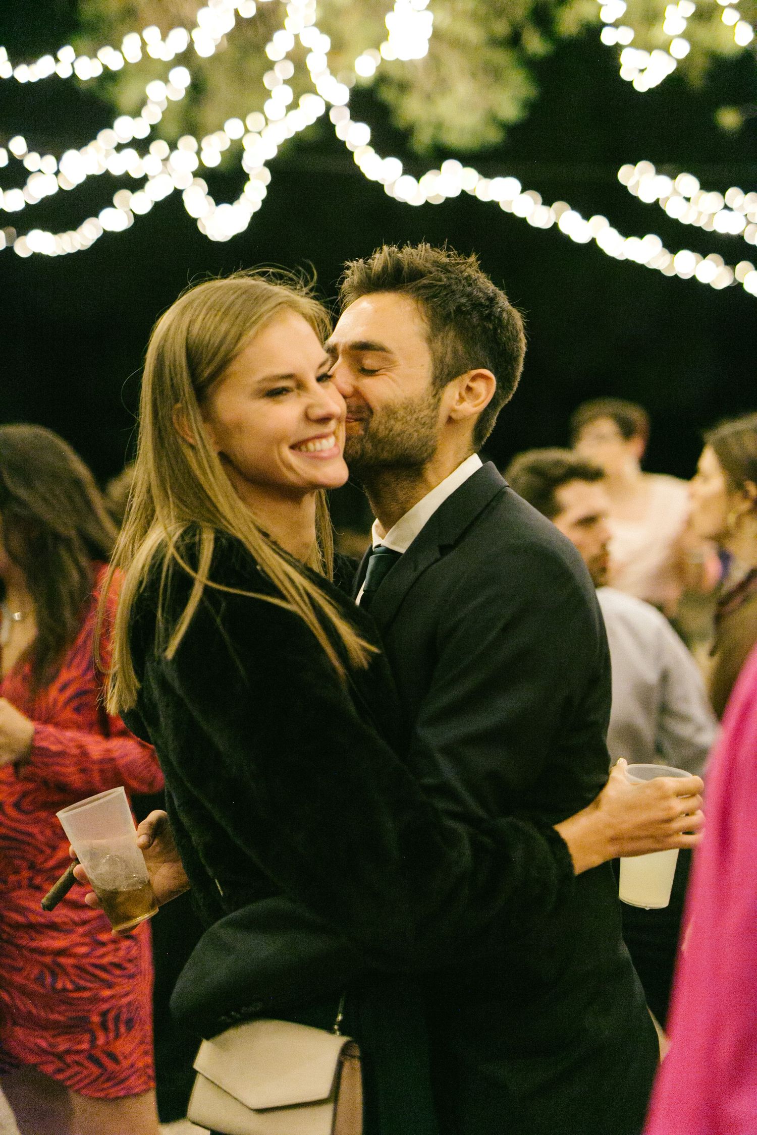 A romantic couple embraces and shares a kiss at an outdoor evening party with string lights glowing in the background.