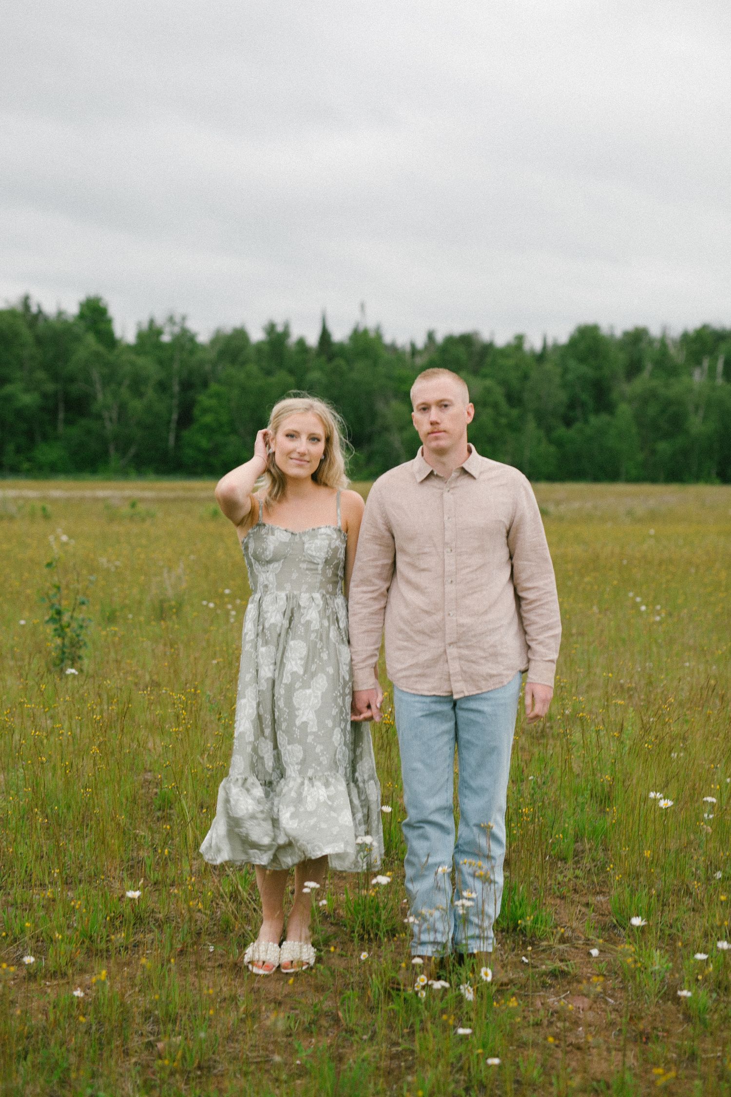 A couple stands together in a wildflower meadow surrounded by trees on an overcast day.