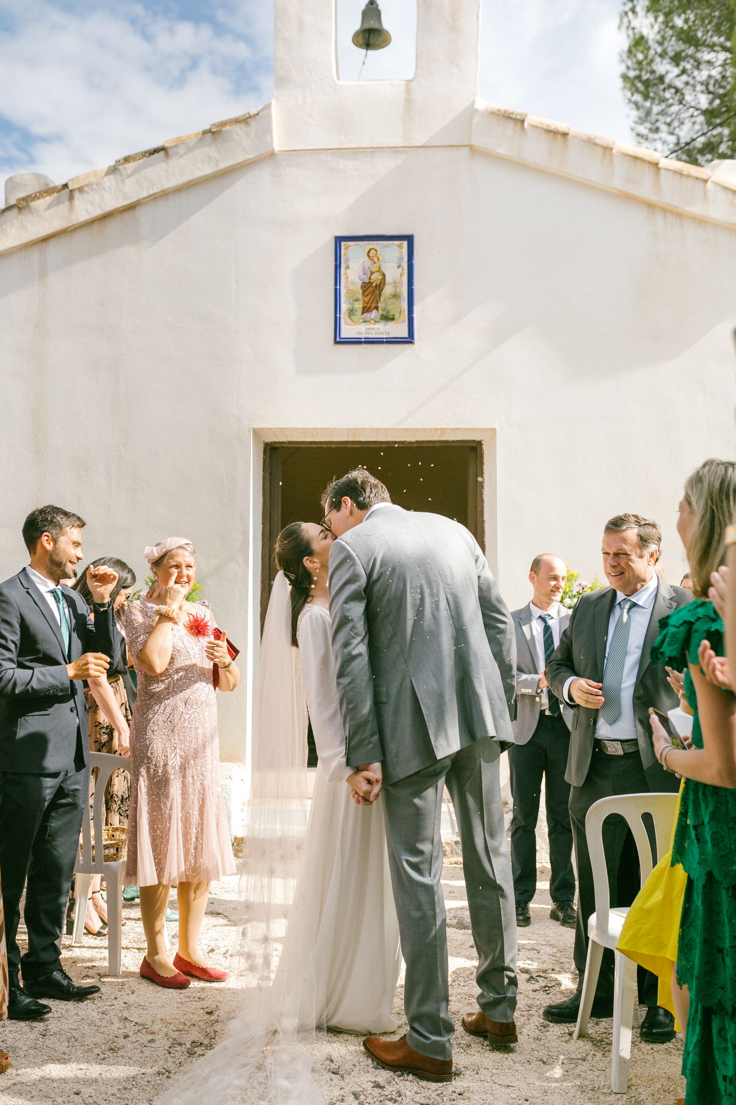 Wedding guests gather outside a small white chapel with a bell tower and religious icon during a sunny celebration.