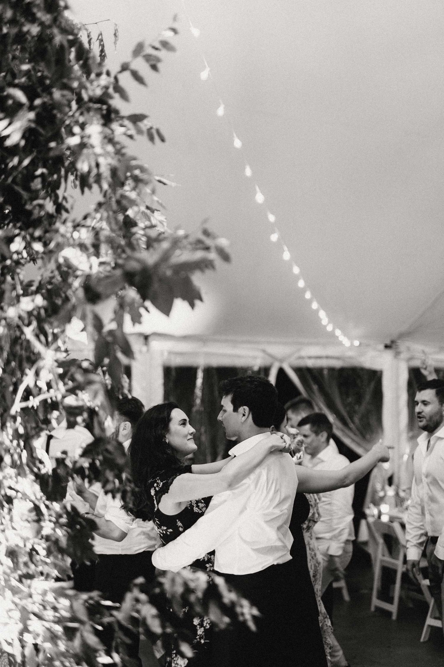 A couple dances together under string lights at an evening wedding reception in a tent.
