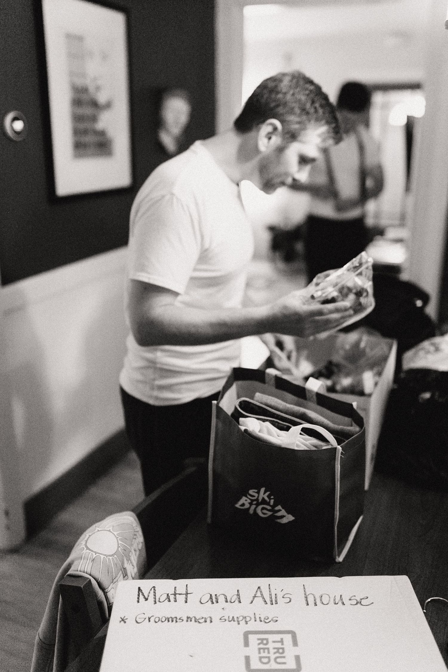 Black and white image of someone in a white t-shirt reading something while standing in a room with a box labeled Matt and Al's house.