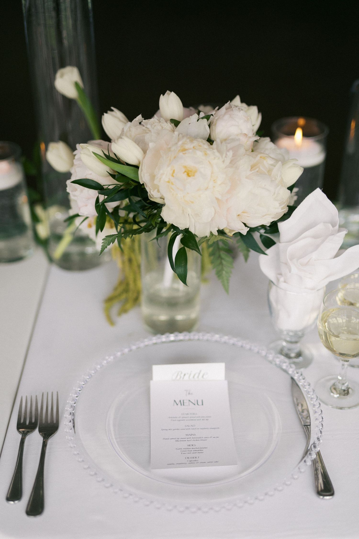 Elegant white table setting with peony centerpiece, crystal plates and formal silverware.