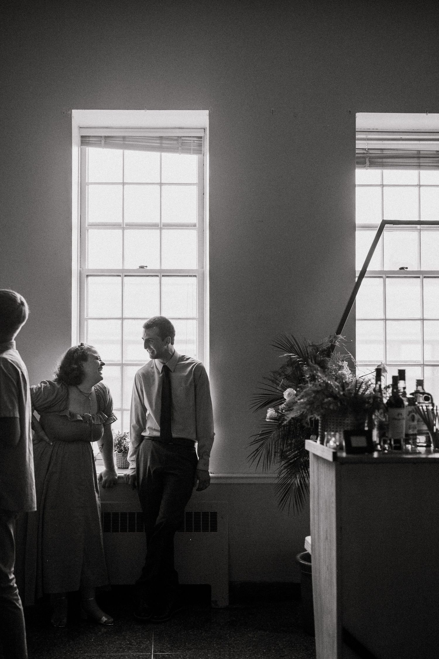 A vintage black and white photo of people gathered by tall windows with a bar cart and palm plant in an interior space.