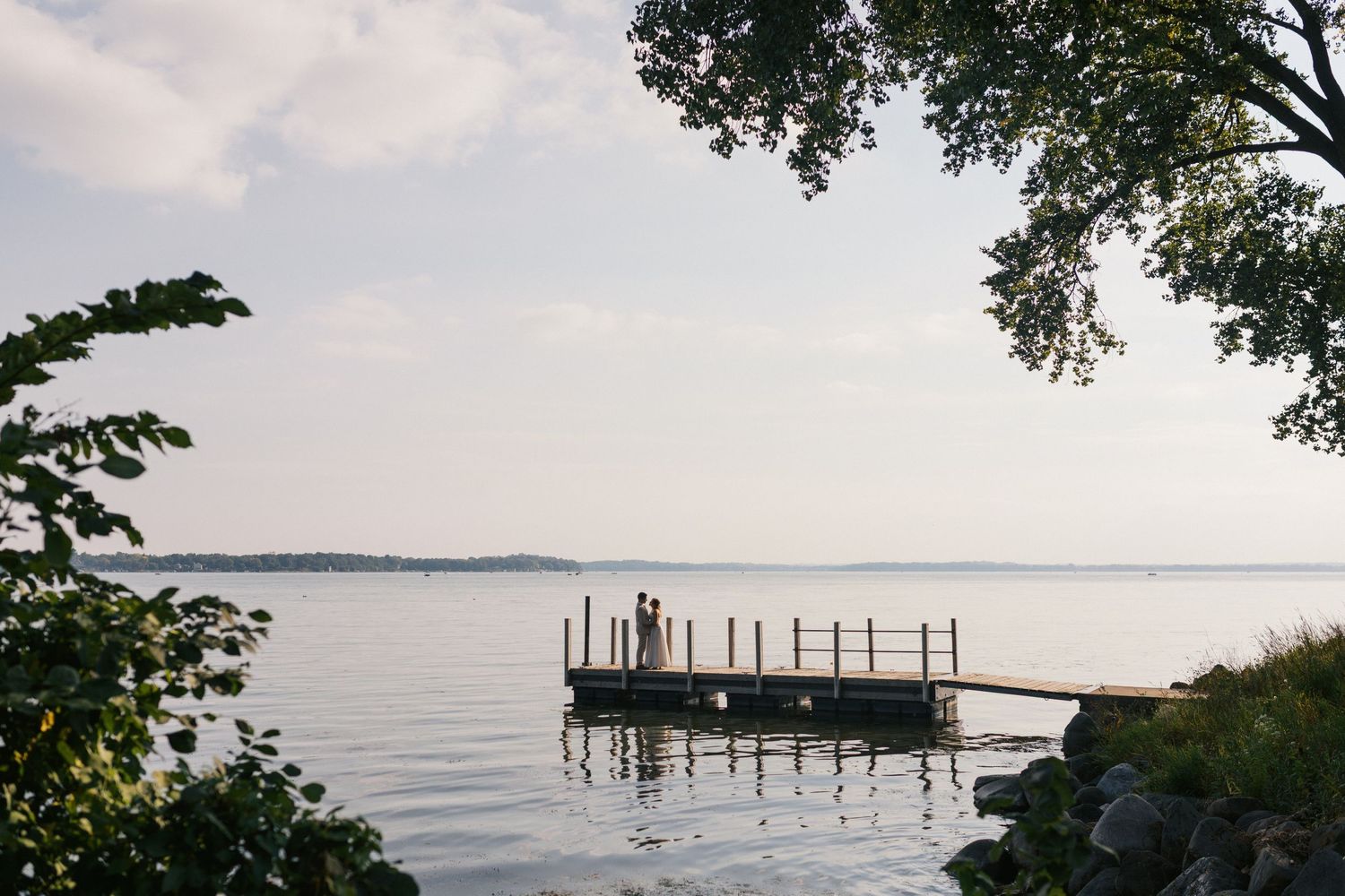 A bride and groom embrace in gentle light on a wooden dock that extends into a calm lake surrounded by trees and rocks at sunset.