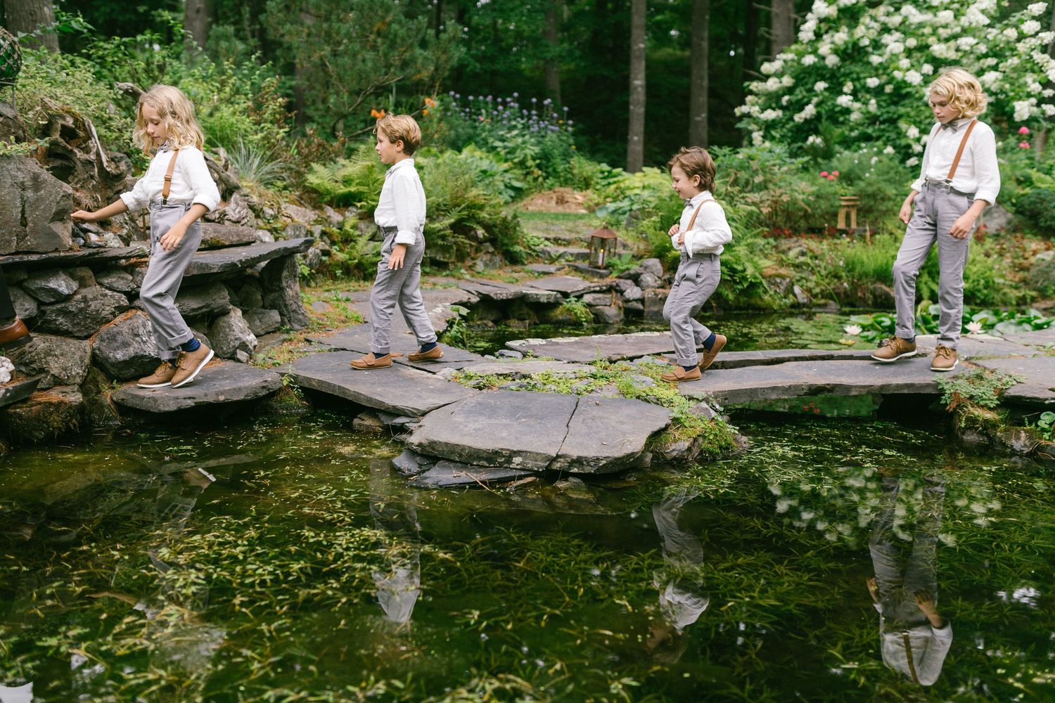 People in white clothing walk along a peaceful garden pond with stepping stones and lush greenery.