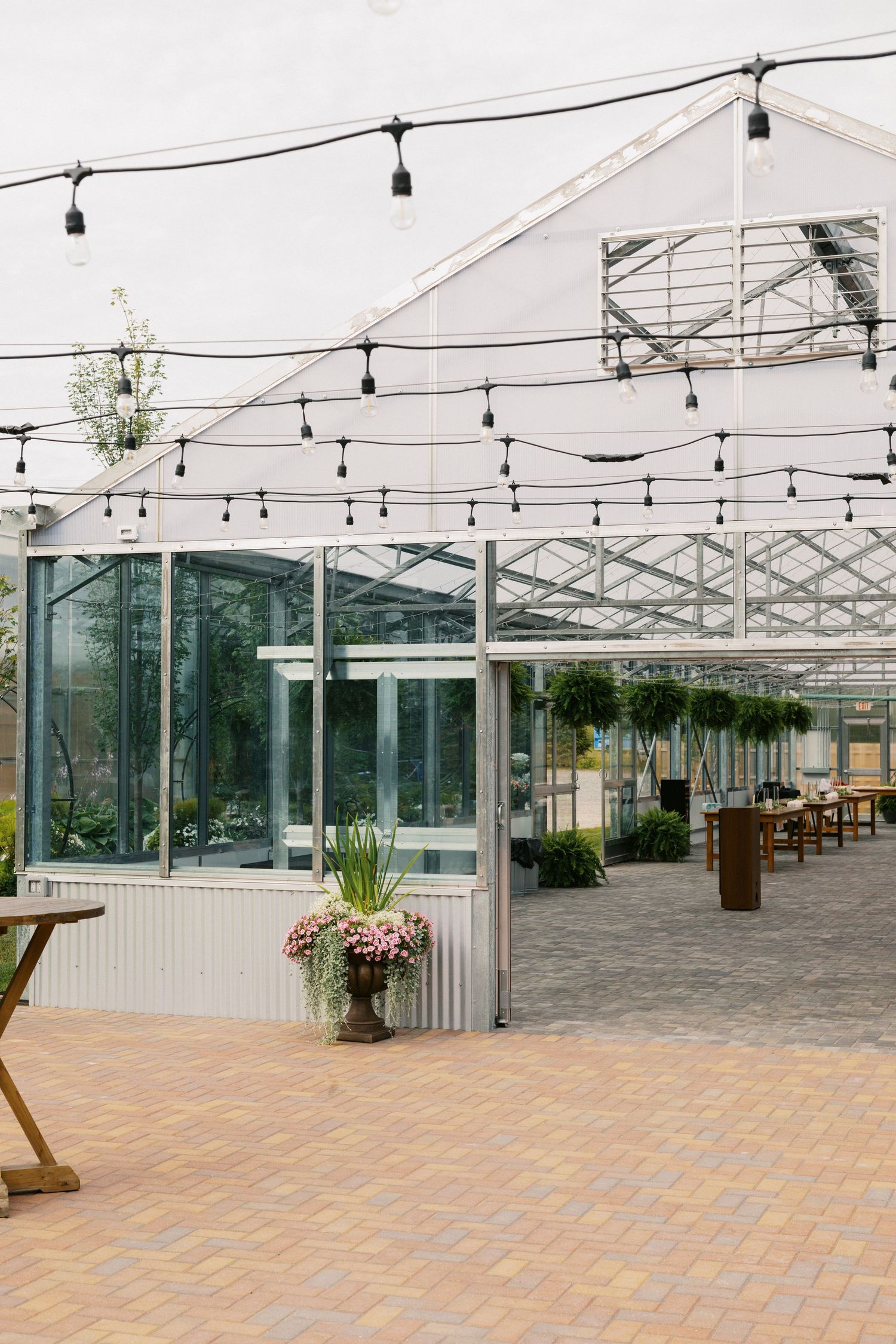 Elegant greenhouse venue with string lights, glass walls, potted flowers, and brick pavers for events.