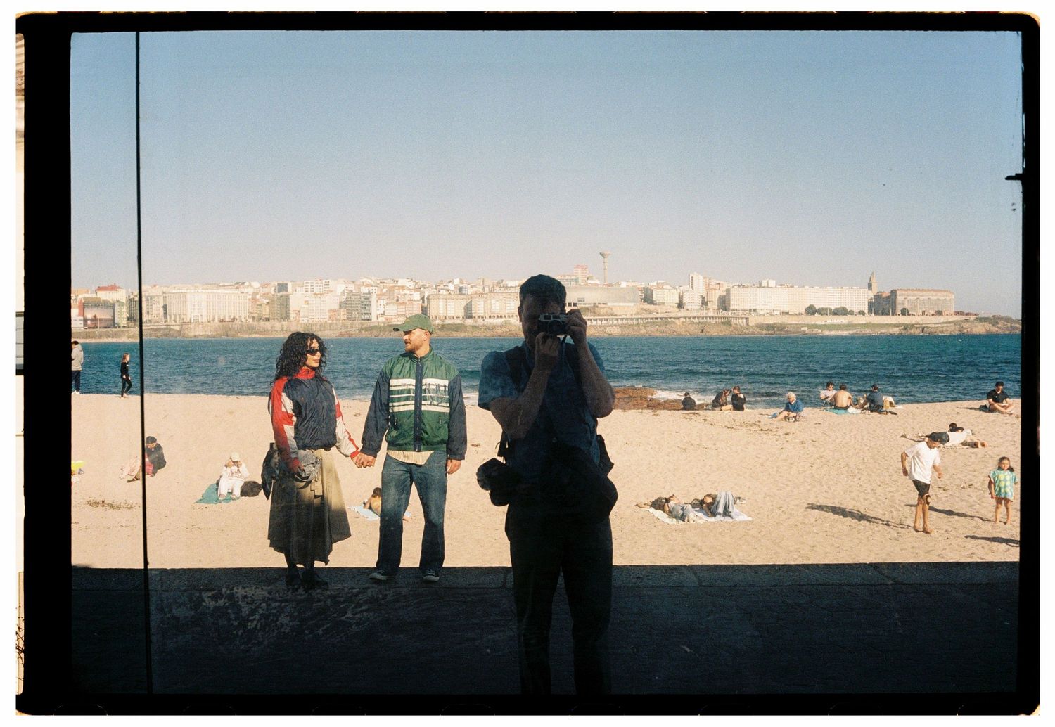 Two people holding hands on a sunny beach with a coastal city skyline in the background.