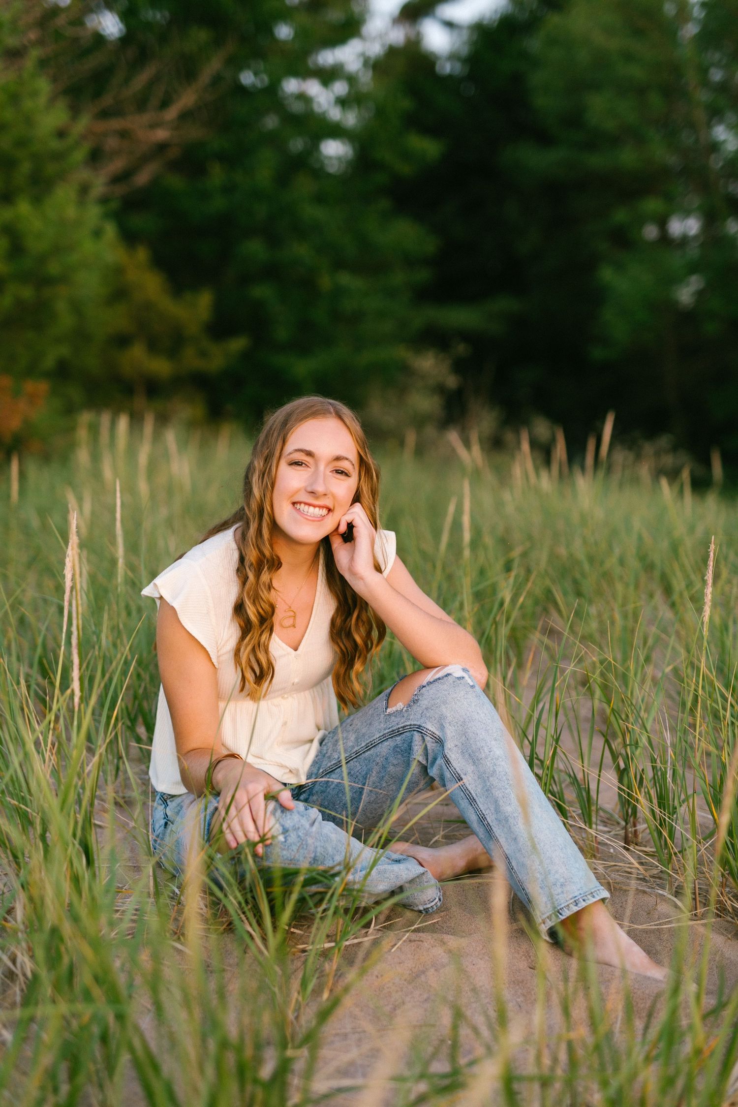 Young woman with wavy hair sits smiling in tall beach grass wearing white top and ripped jeans at golden hour.