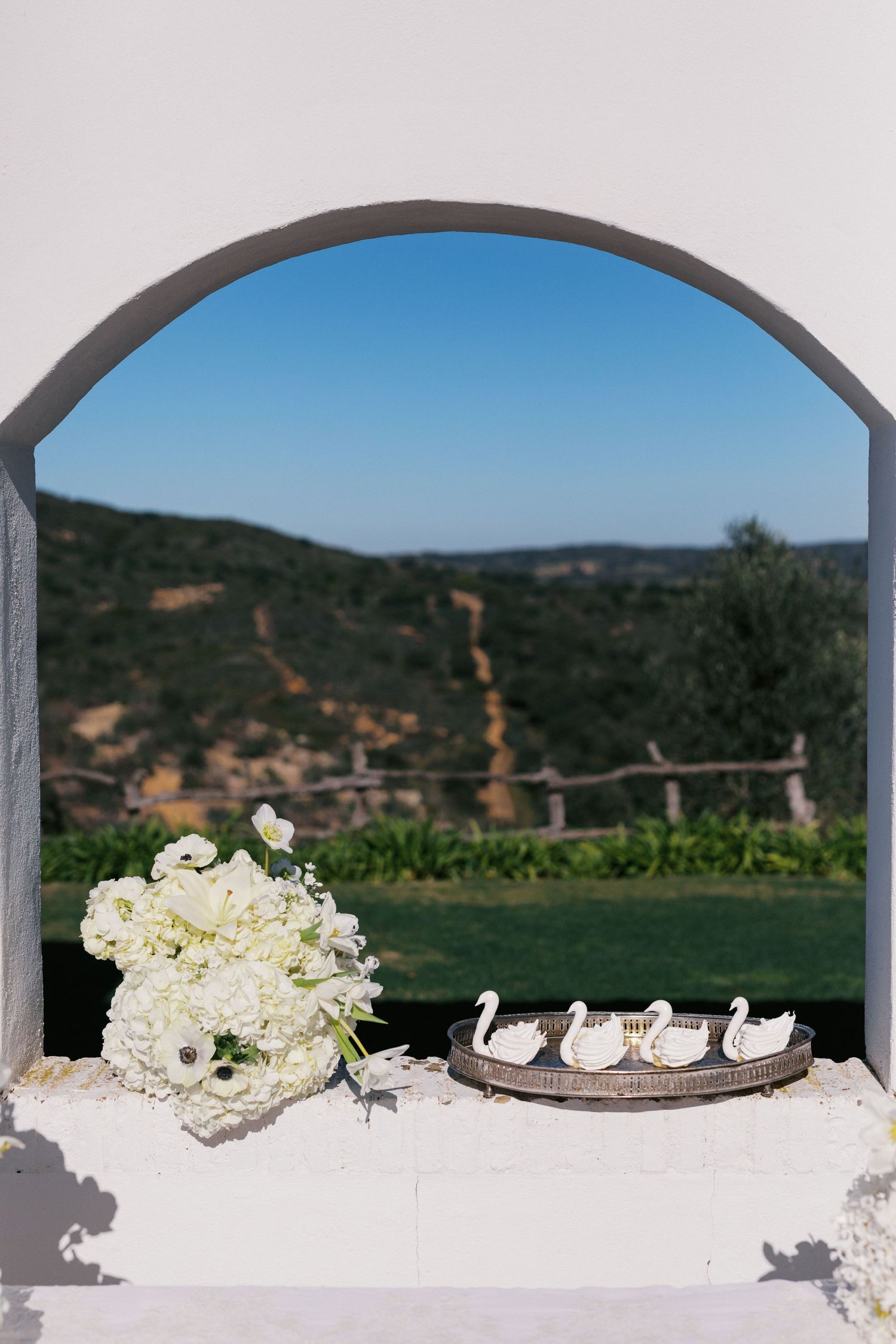 White floral bouquet and swan figurines on a table framed by an arch with scenic green hills backdrop.