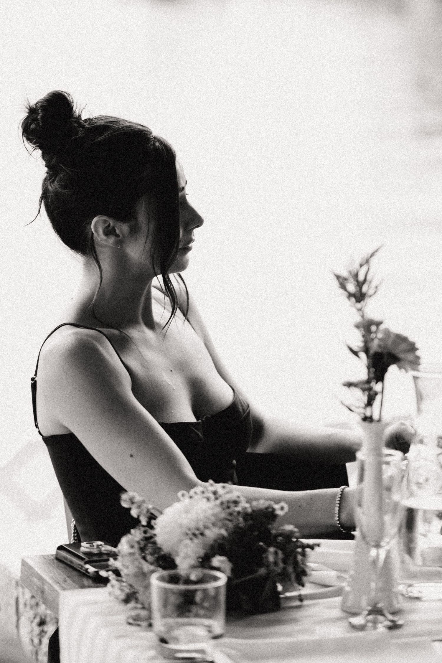 Black and white portrait of a figure with hair in a bun sitting at a table with flowers in vases.
