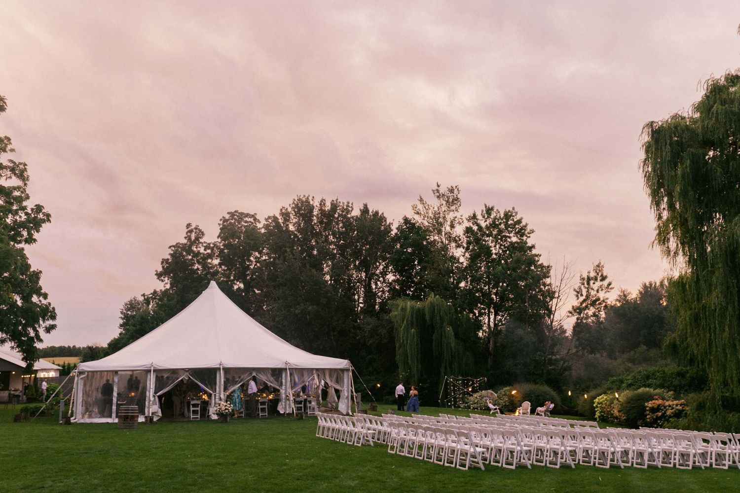 A panoramic view of an outdoor wedding venue with white chairs set up on a green lawn under a cloudy sky.
