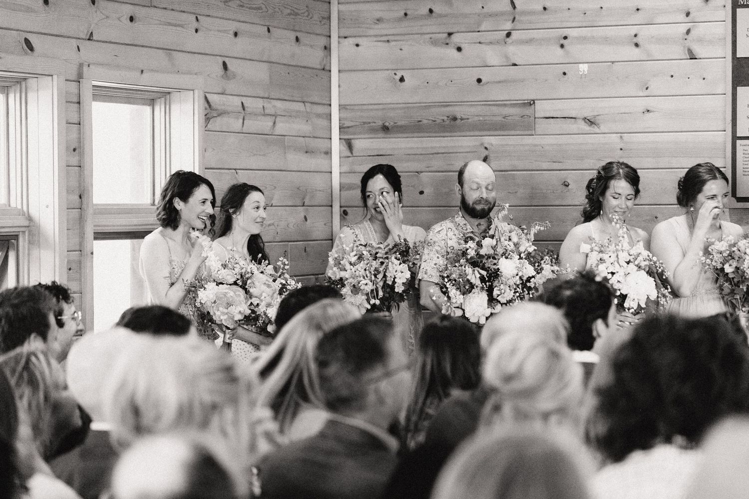 A black and white photograph shows a wedding party lined up at the front of a rustic wooden room filled with guests.