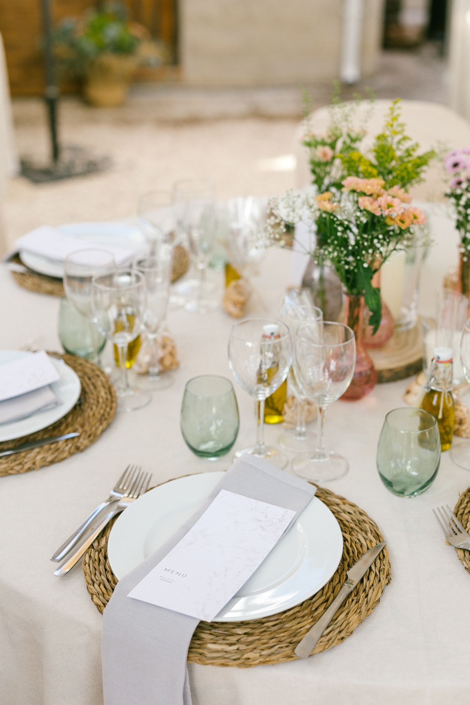 Elegant table setting with woven placemats, white plates, glassware and floral centerpieces on cream tablecloth.