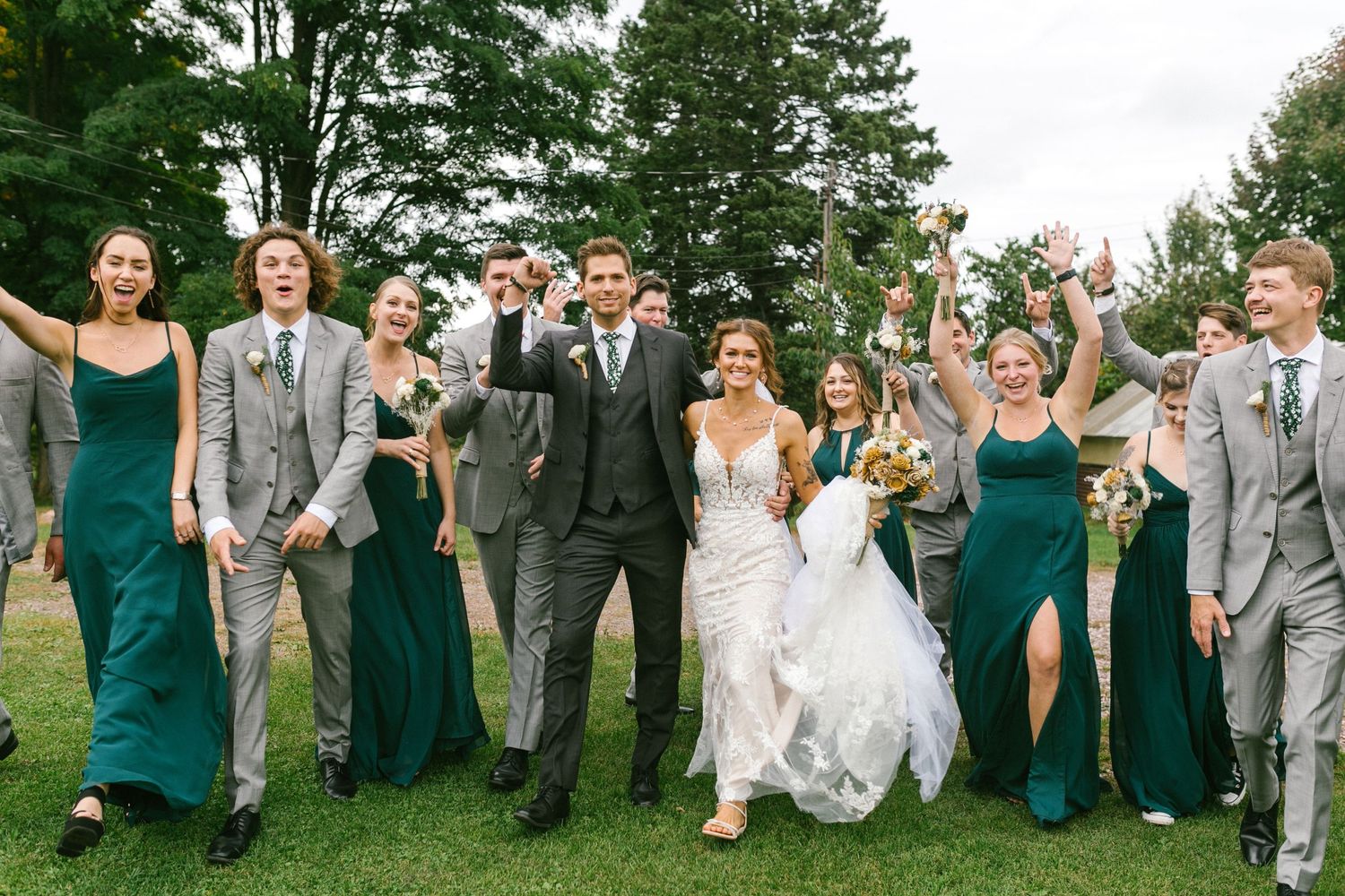 Wedding party in emerald green dresses and gray suits walking and celebrating joyfully on grassy lawn.