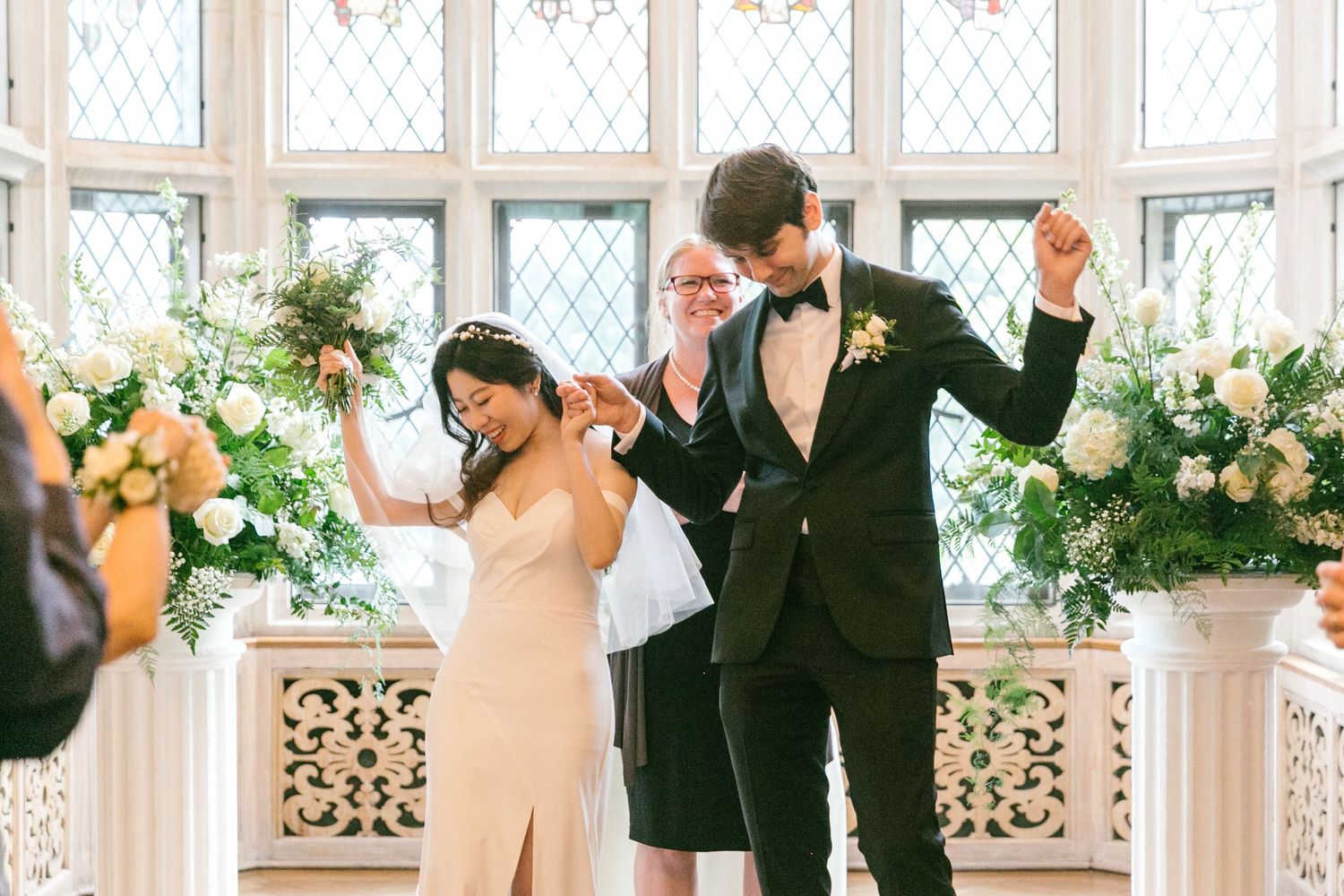 Newlyweds celebrate with raised hands at altar decorated with lush white floral arrangements in bright chapel.