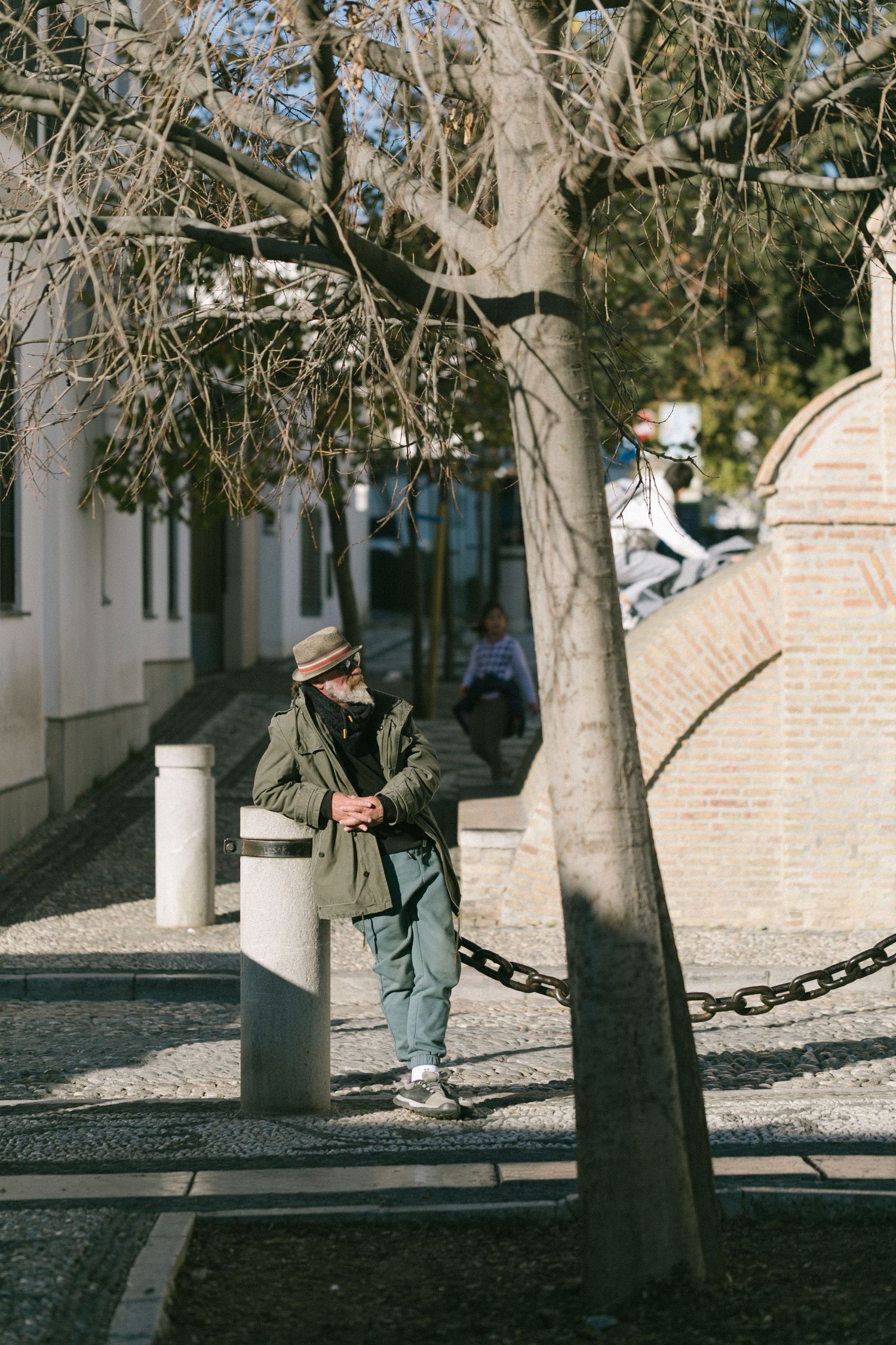 Person stands by a tree on a cobblestone street with white buildings and bollards in an urban setting.