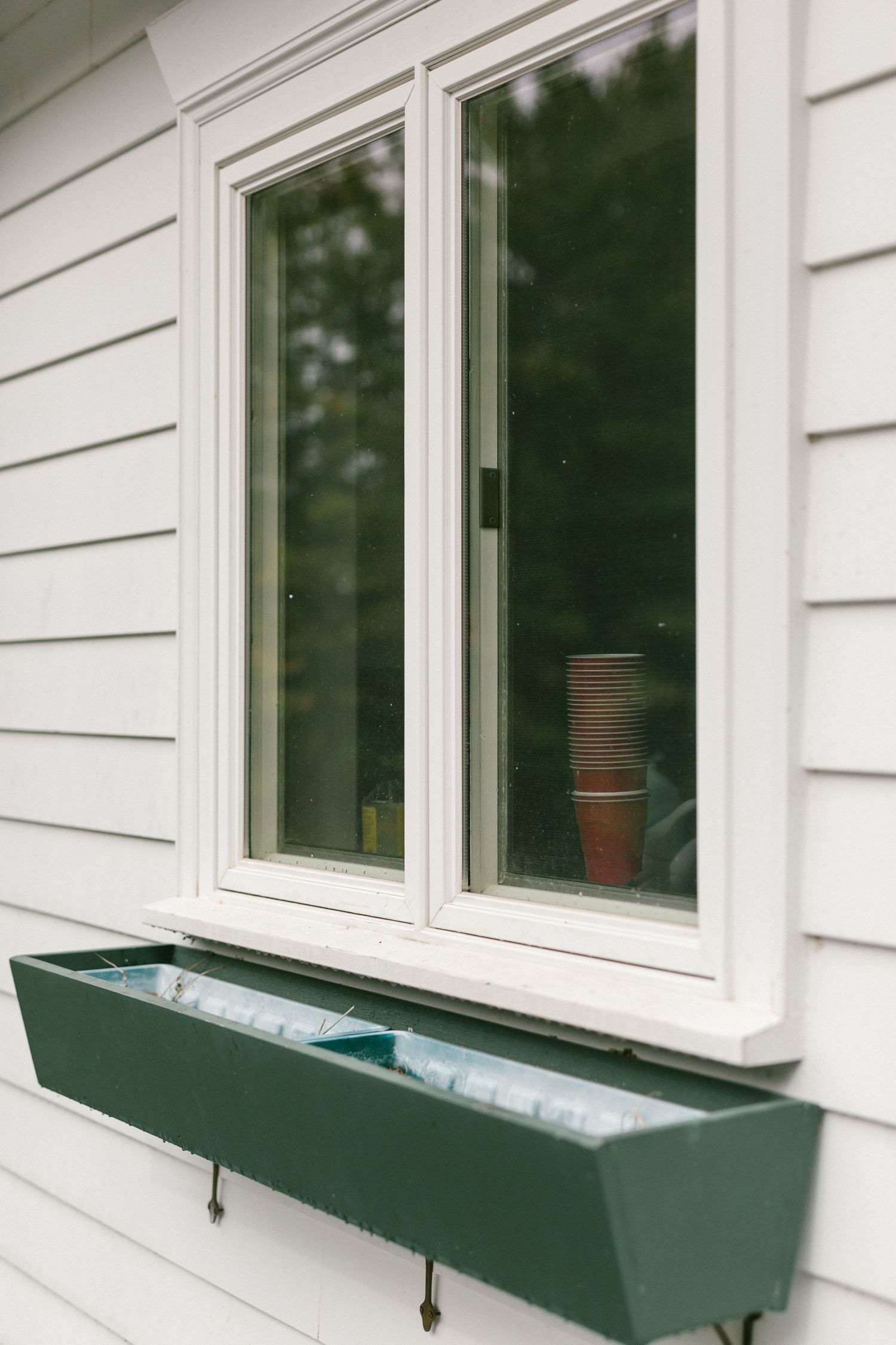 A dark green window box planter mounted beneath white vinyl siding and double-pane windows of a house.