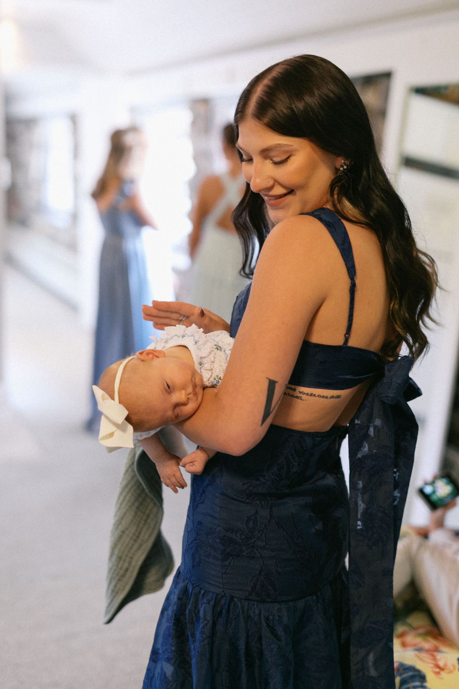 A woman in a navy blue dress holds a small baby in an indoor setting.