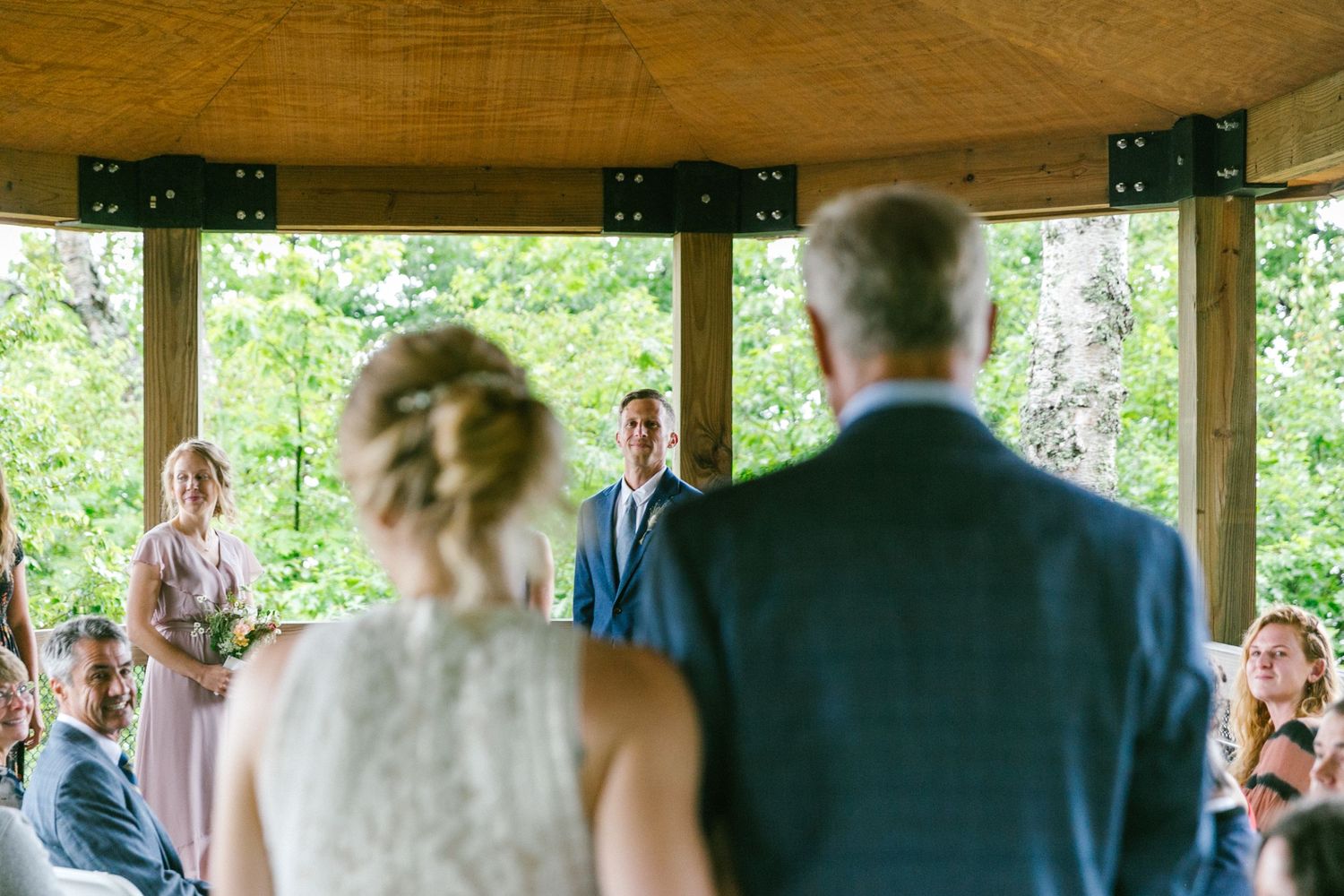 Intimate outdoor wedding ceremony in a wooden pavilion with natural forest backdrop.