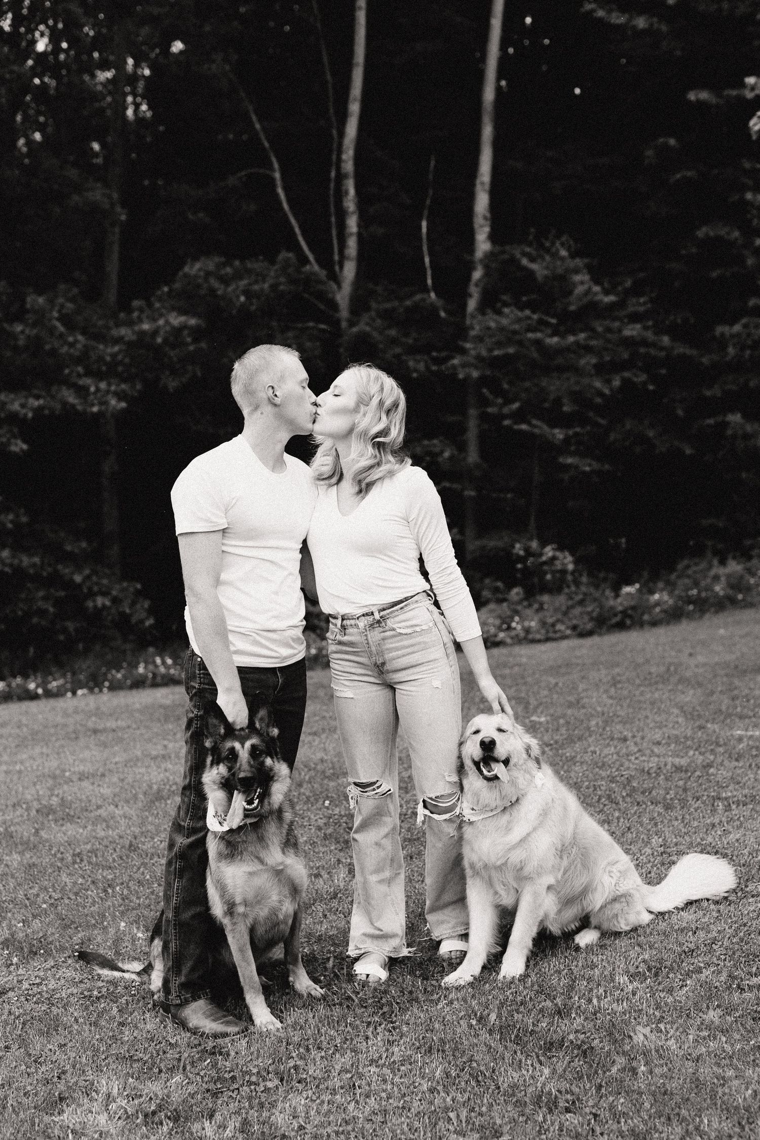 A couple shares a tender moment outdoors with their two dogs, a German Shepherd and Golden Retriever, in a black and white photo.