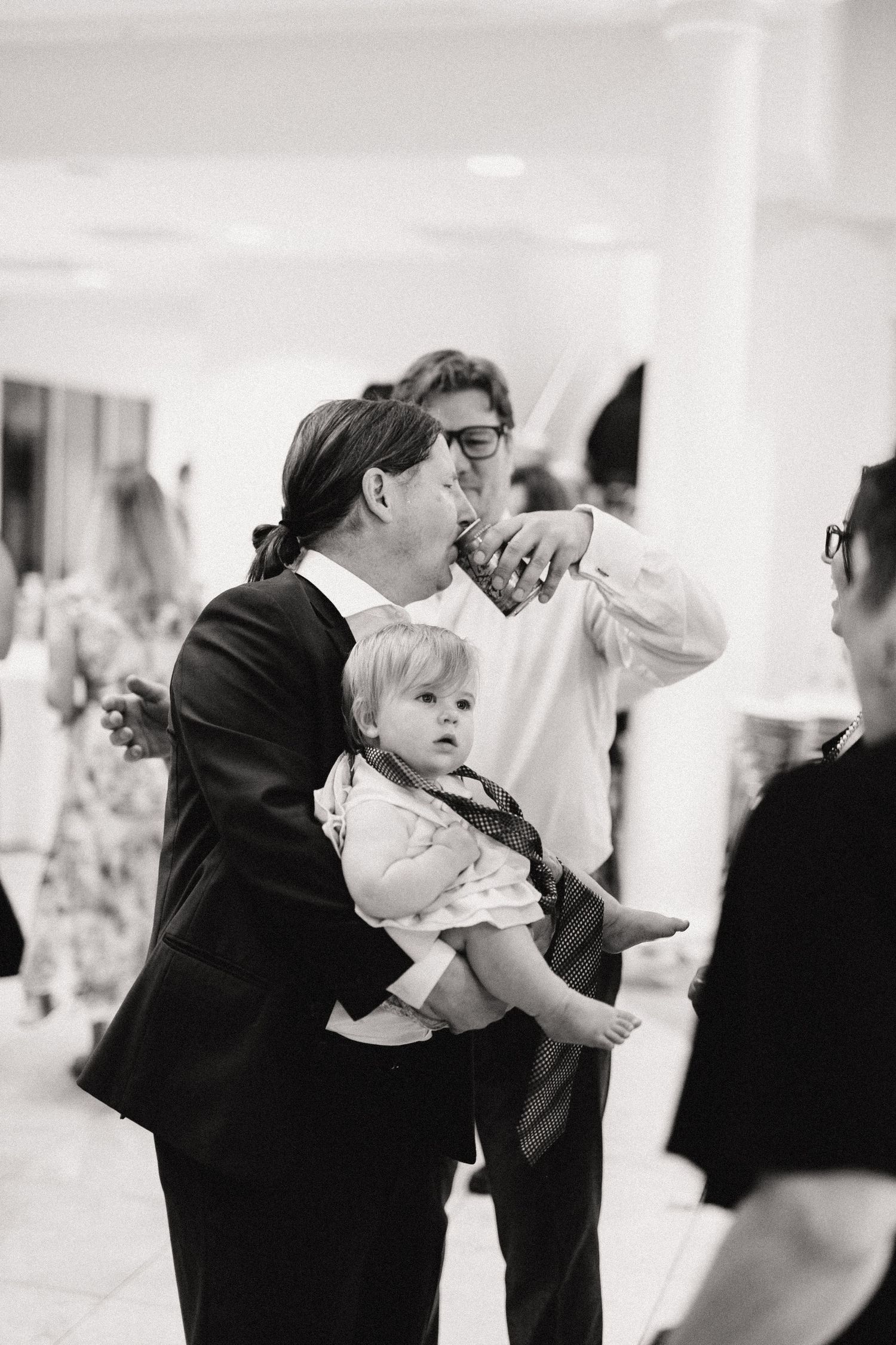 A man in a suit holds a small child in formal wear at what appears to be an indoor event, captured in black and white.