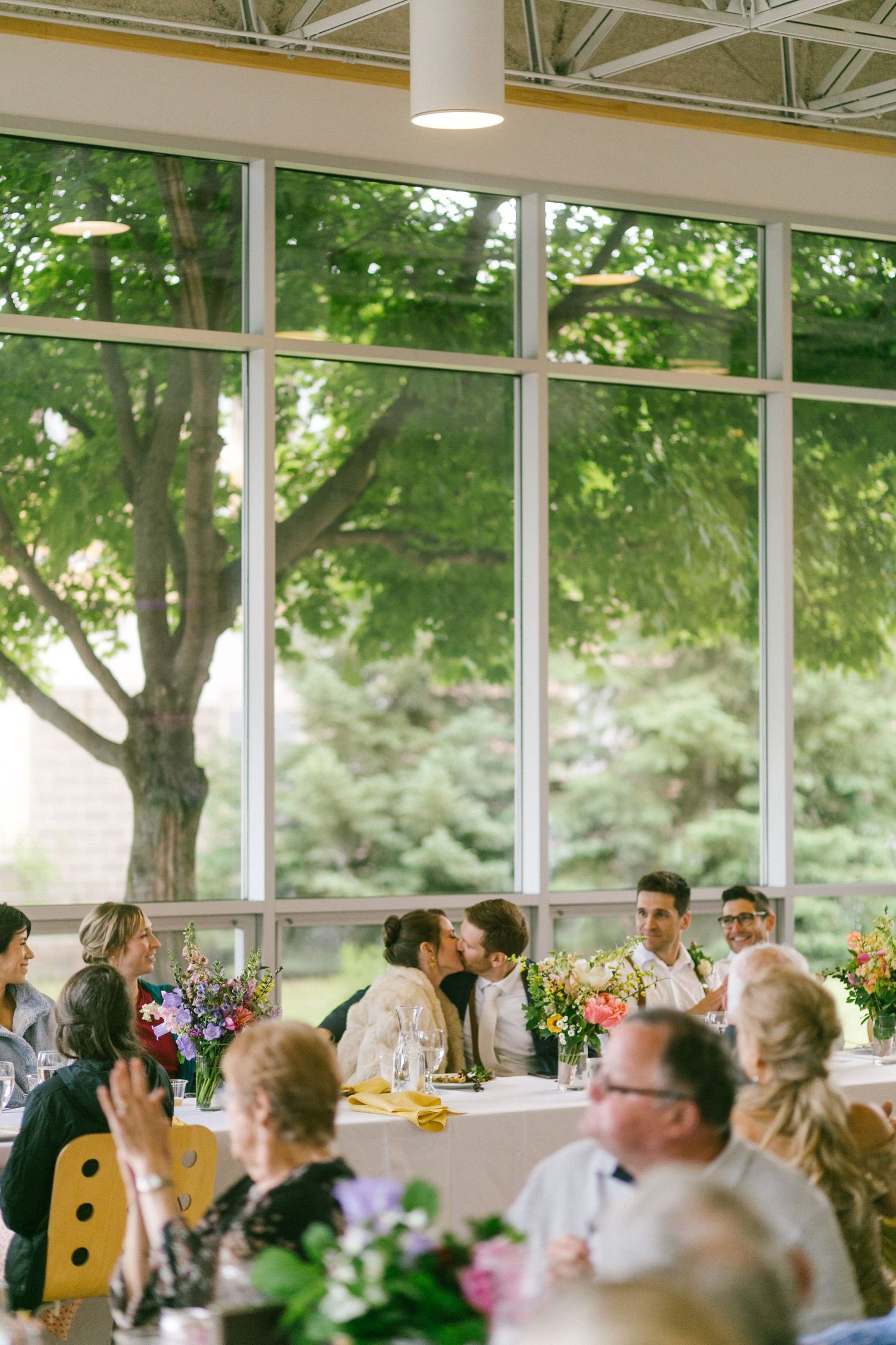Guests gather at elegant reception tables with floral centerpieces in bright sunlit room with floor-to-ceiling windows overlooking trees.