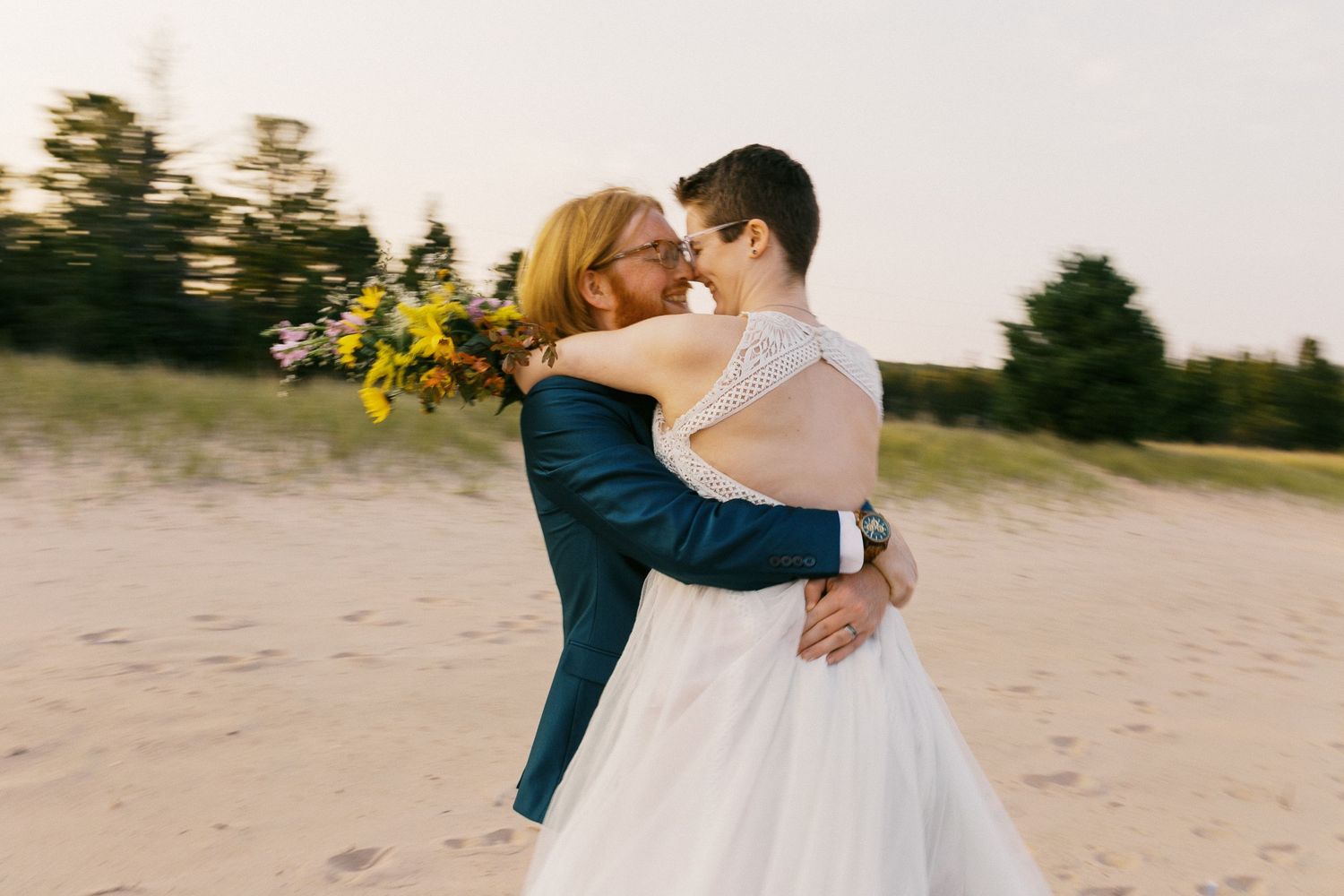 Bride and partner share romantic embrace on sandy beach at golden sunset.