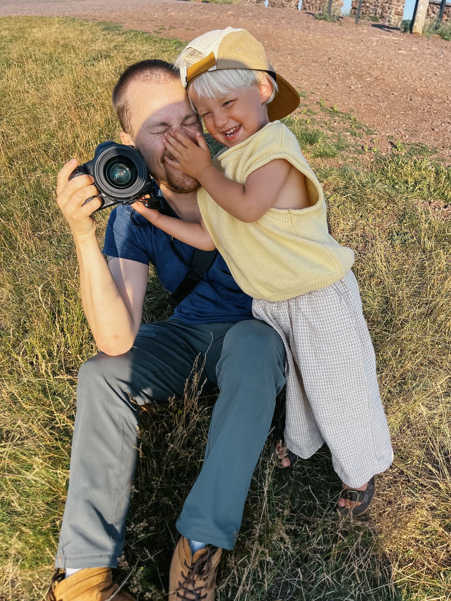 Man photographing with DSLR camera while laughing child in yellow hat leans on him outdoors.