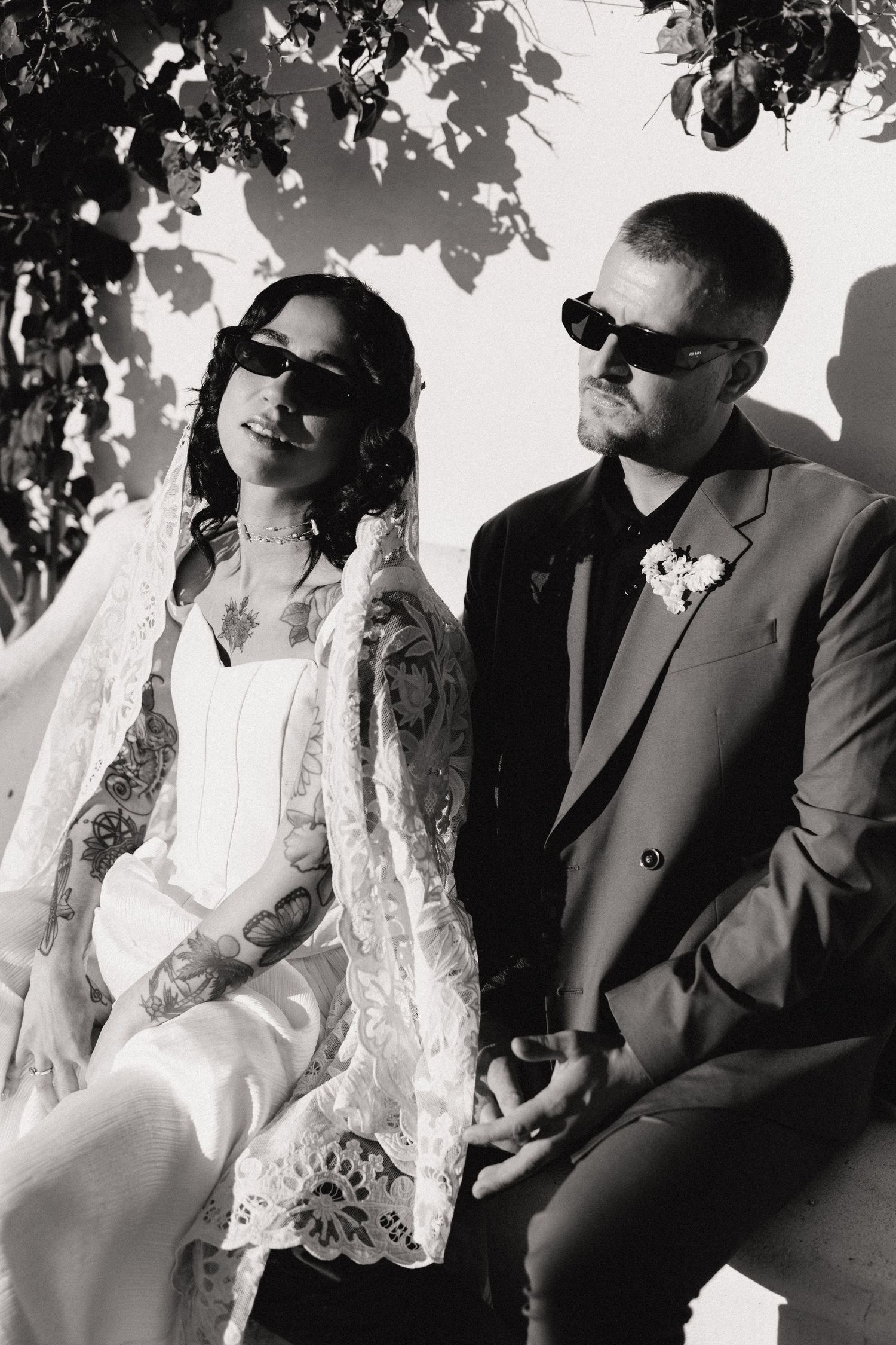 Black and white photo of tattooed bride and groom wearing sunglasses, bride in lace veil, groom in dark suit.