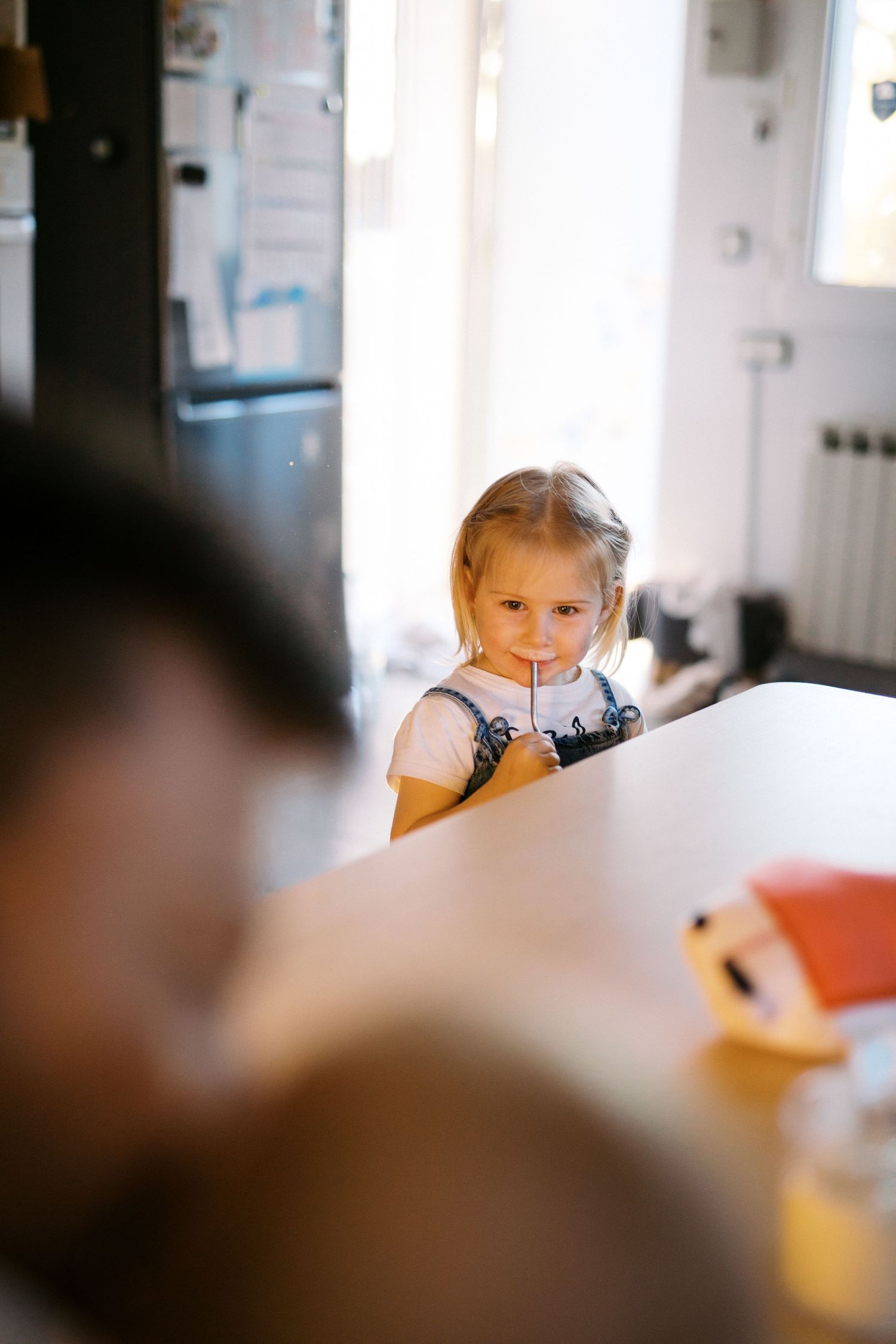 Young blonde girl drinking through a straw at a kitchen table, looking at camera in bright home interior.