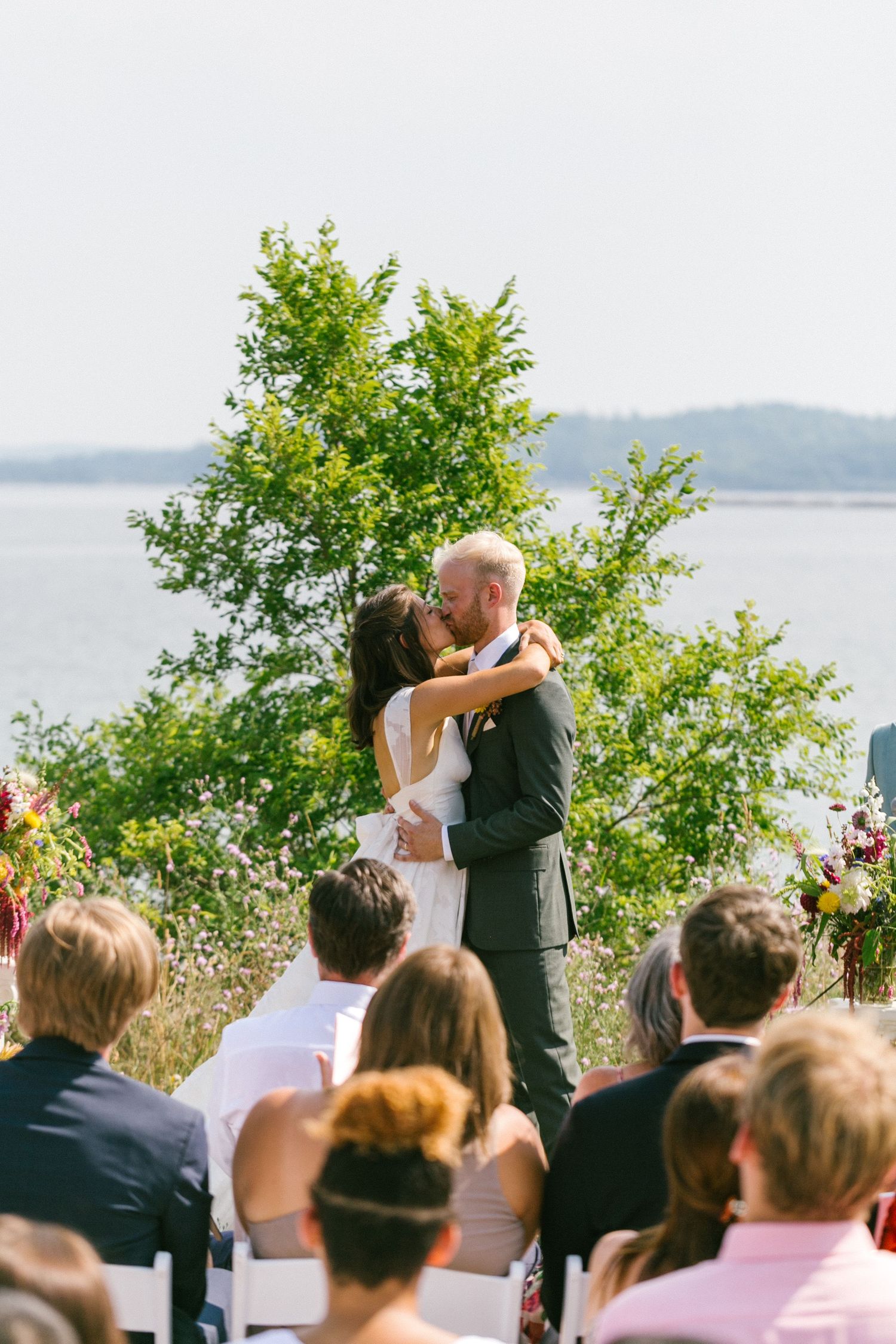 An outdoor wedding ceremony takes place with guests watching by the waterfront.