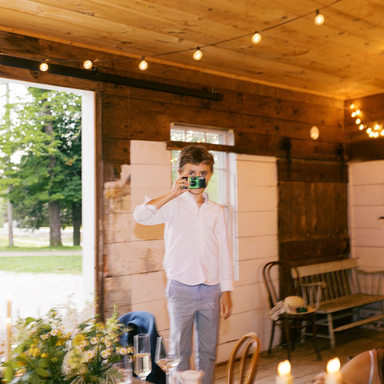 A young man photographs with a green camera inside a rustic barn decorated with string lights at a wedding reception.