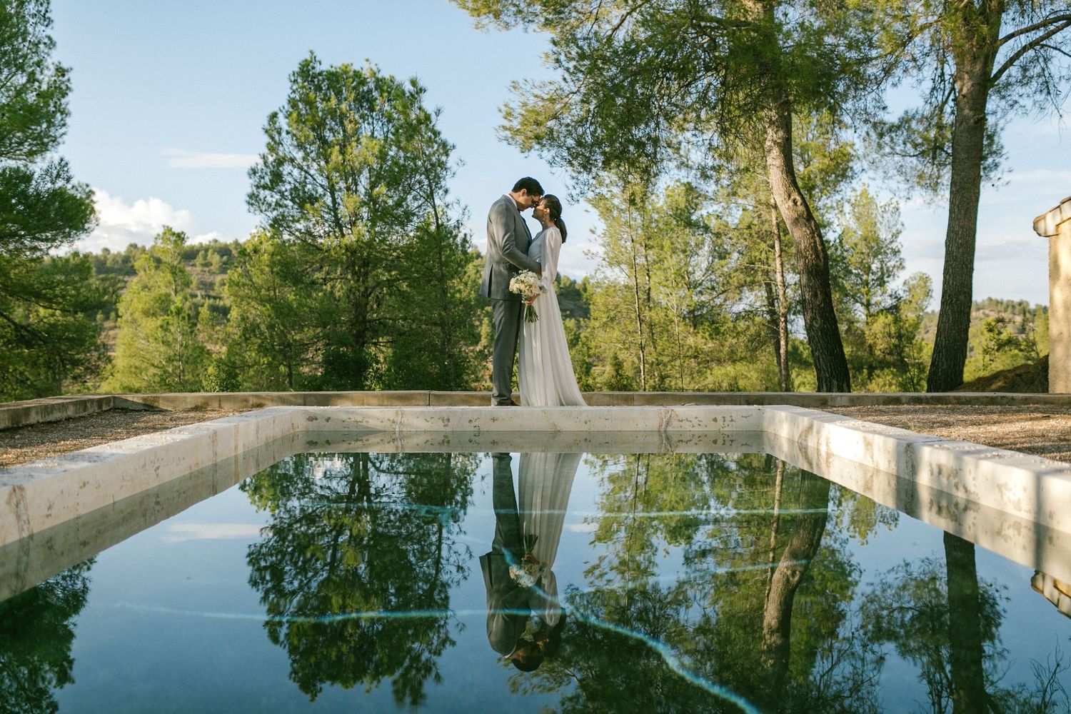 A couple stands embracing beside a reflective pool surrounded by trees in a serene outdoor setting. Europe destination wedding photographer.
